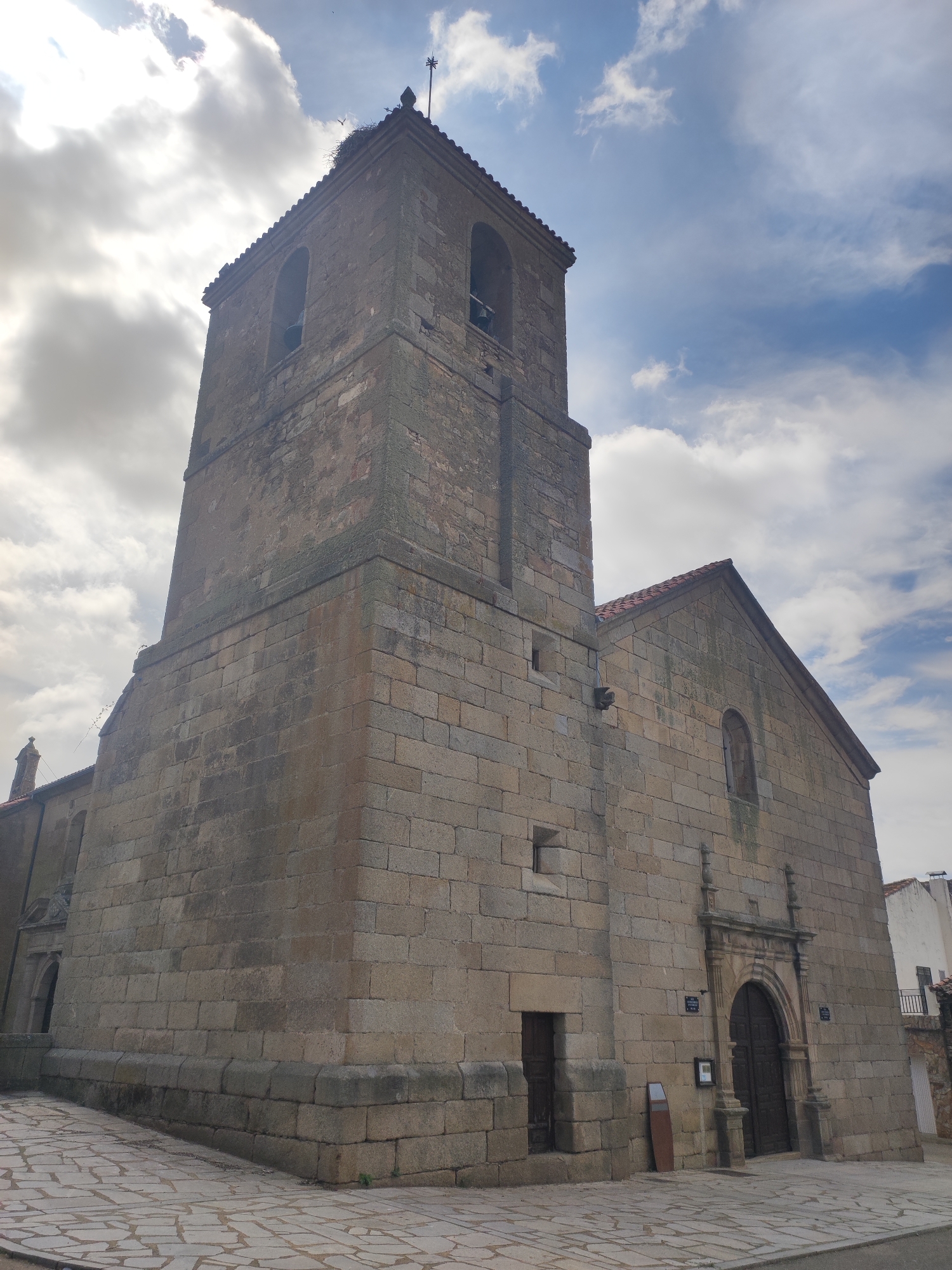 The image shows a stone church in Sobradillo, Spain. The church has a bell tower with arched openings, and a steeple with a bird's nest at the top. The main body of the church is constructed from large, rough-hewn stones. A large wooden door is visible on the facade, which is also made of stone. The ground in front of the church is paved with irregular flagstones. The sky is partly cloudy, with patches of blue and white clouds.