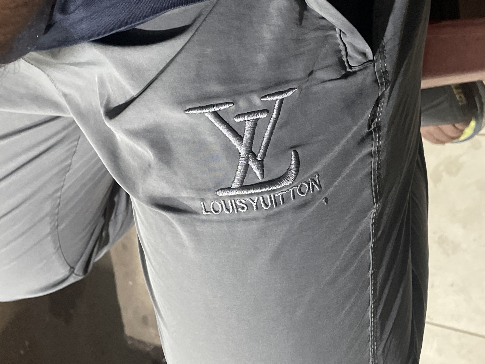 A close-up shot depicts gray Louis Vuitton track pants, showcasing the embroidered LV logo and brand name. The location is unknown; no information indicates the scene is in Dhaka, Bangladesh. A fragment of a blue shirt is visible at the top of the image, and a portion of a shoe is visible in the background.