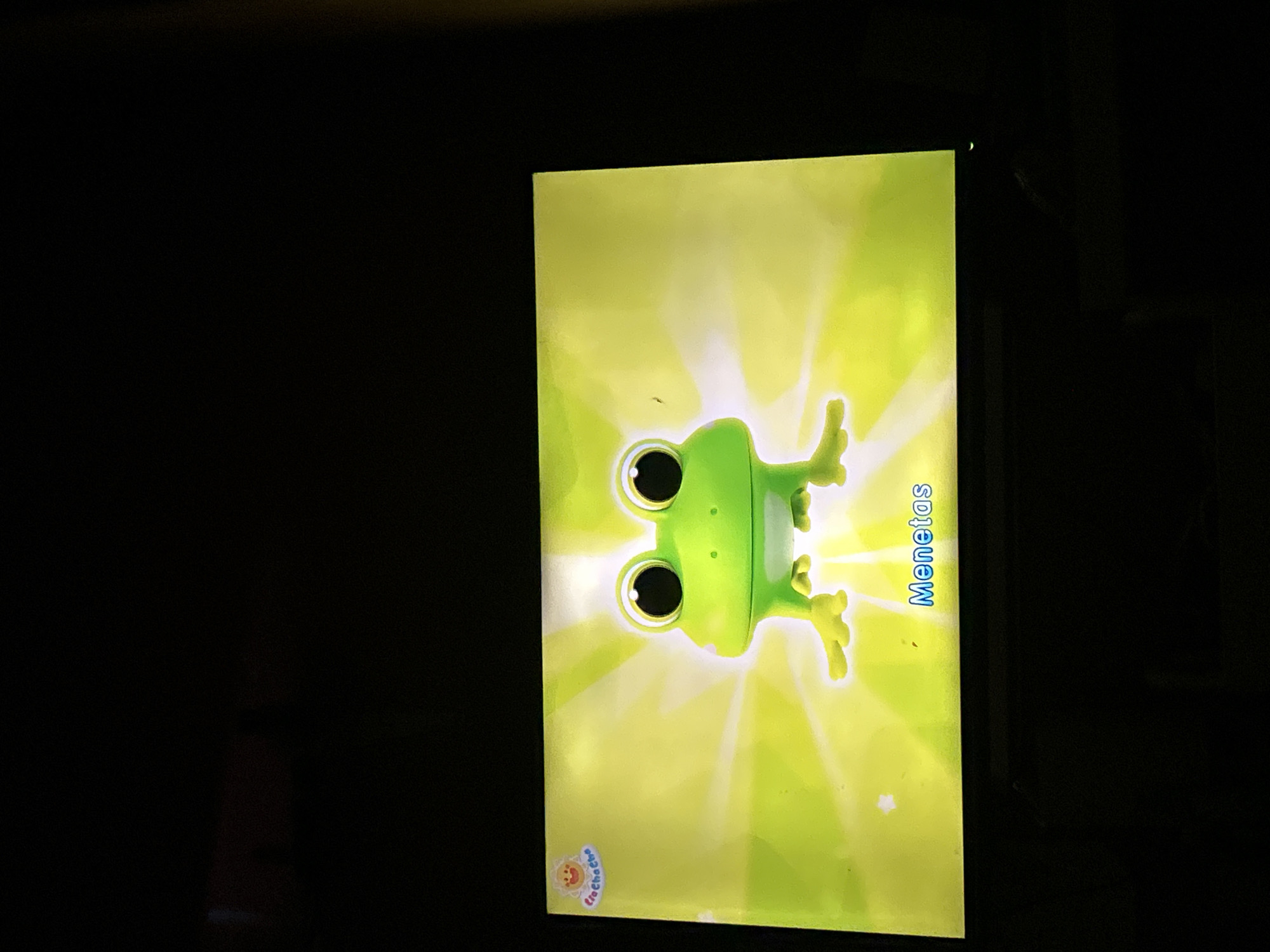 A 3D animated green frog with large, black-eyed eyes and a wide smile is centered on a bright yellow background that emits light rays. The frog is facing forward and has its front legs splayed out. In the top left corner, there is a small circular logo with a smiling sun and text that reads "CioChaCho." In the bottom right of the screen, the word "Menetas" is written in blue text. The image appears to be a screenshot from a children's educational program or animation. The scene is not identifiable as being in Depok, Indonesia.