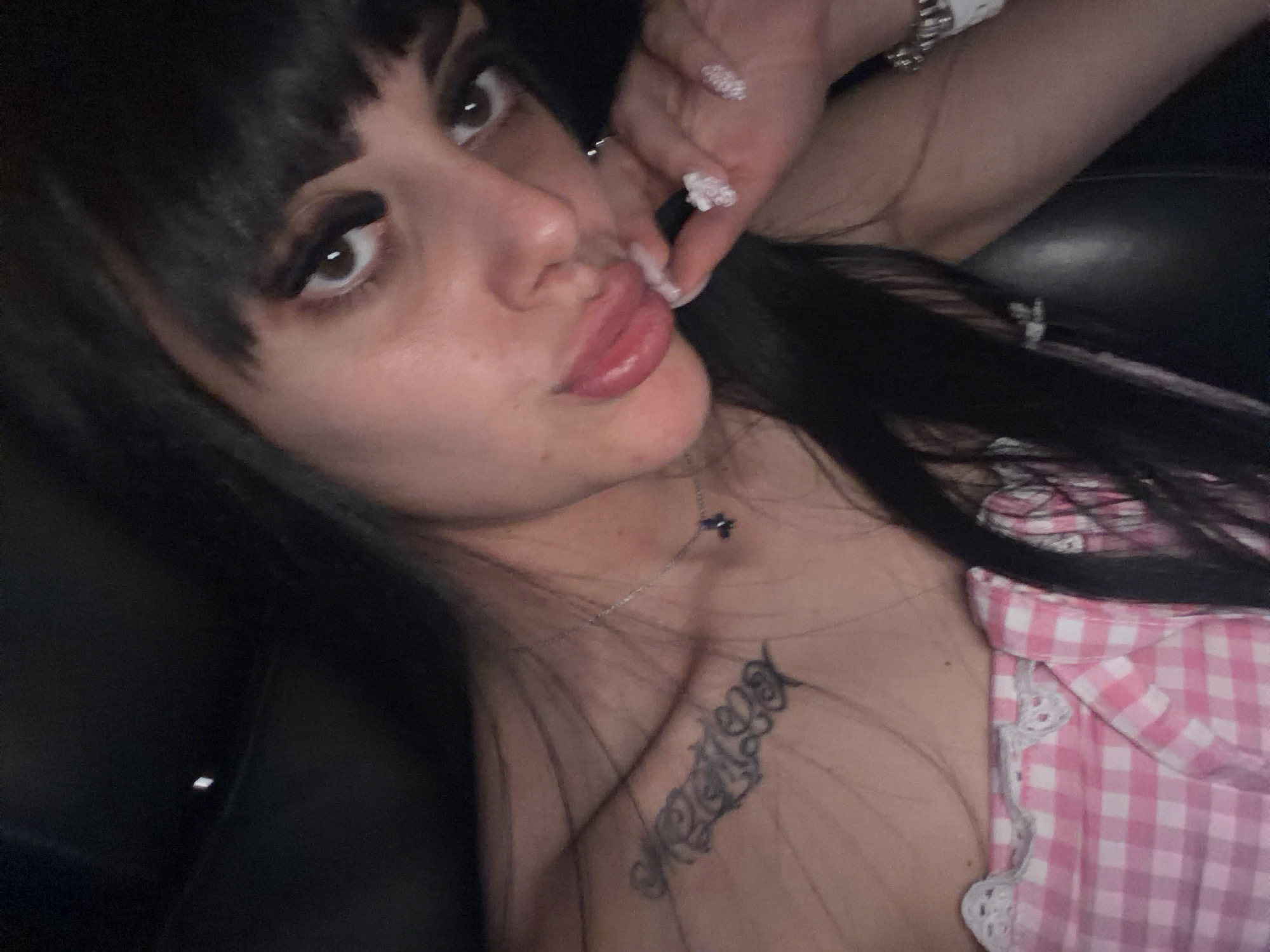 The image shows a close-up, self-portrait style photograph of a young woman with dark hair and bangs. She is wearing a pink and white checkered top with lace trim. Her left shoulder is visible, revealing a tattoo on her collarbone and a delicate silver necklace with a dark blue pendant. Her right hand is near her face, and her nails are decorated with white glitter. She is looking at the camera with a slightly parted mouth. The background is dark and appears to be indoors, possibly a car interior, given the dark, slightly reflective surface. There are no discernible landmarks or features that would indicate a specific location within Plovdiv, Bulgaria.