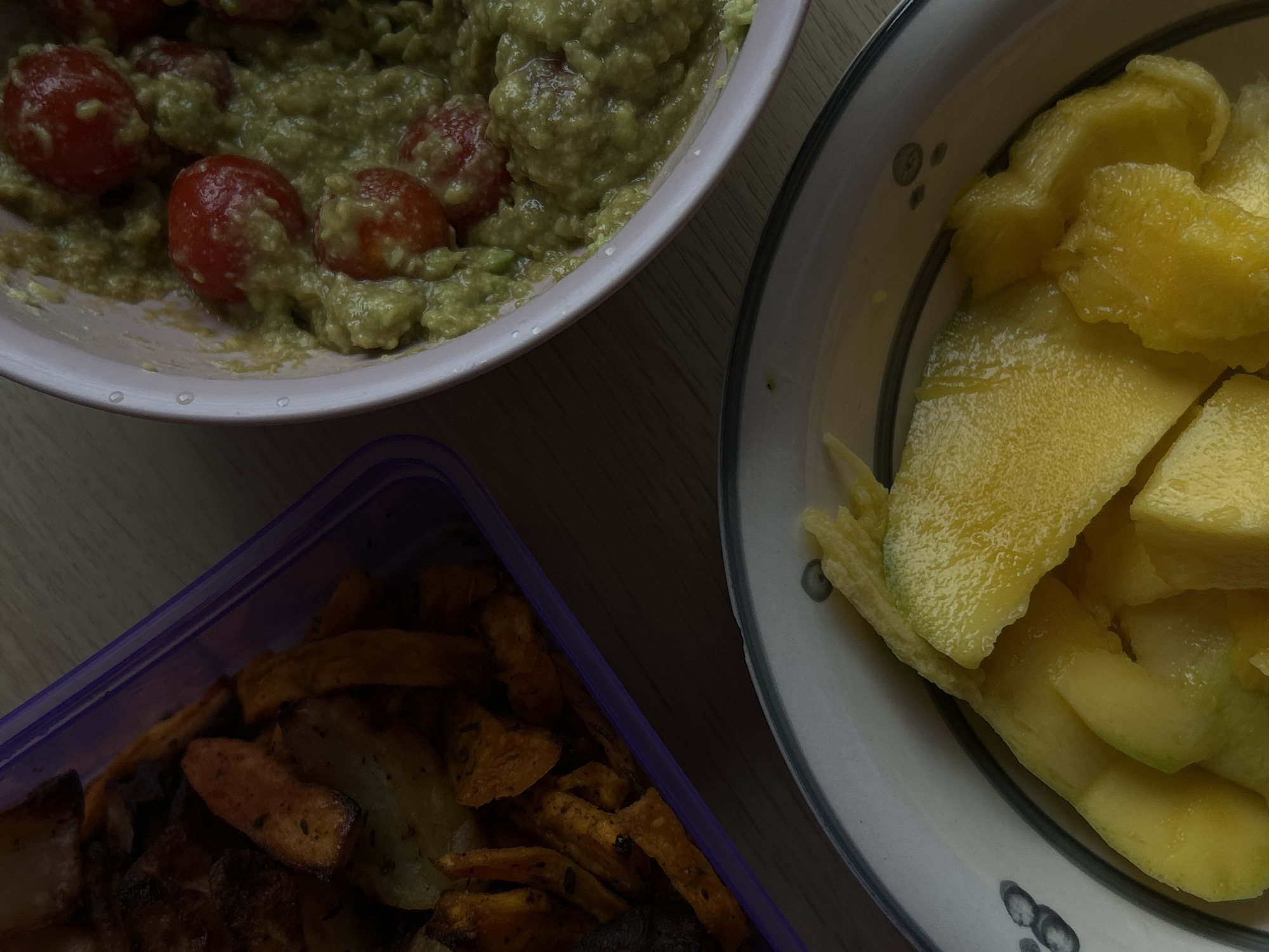 The image shows a top-down view of a meal laid out on a wooden-patterned surface. The meal consists of three dishes. In the top left, there's a pink bowl containing guacamole with cherry tomatoes mixed in. To the right, a plate with sliced mango is visible. At the bottom left, there is a plastic container with what appears to be roasted potato wedges or fries. The dishes are arranged somewhat haphazardly on the surface, suggesting a casual setting. There is no evidence to suggest this picture was taken in Sofia, Bulgaria.