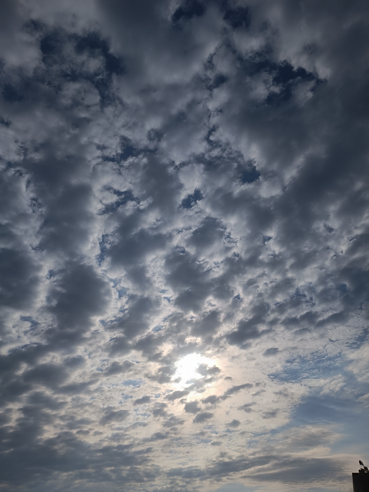 The image depicts a sky filled with a mix of dark and light gray clouds. The sun is partially visible behind the clouds, creating a bright glow. The clouds appear dense and textured, with some areas allowing glimpses of blue sky. The bottom right corner of the image shows a sliver of a dark structure, possibly a building, with some small objects on top. The overall impression is of an overcast sky with diffused sunlight.
