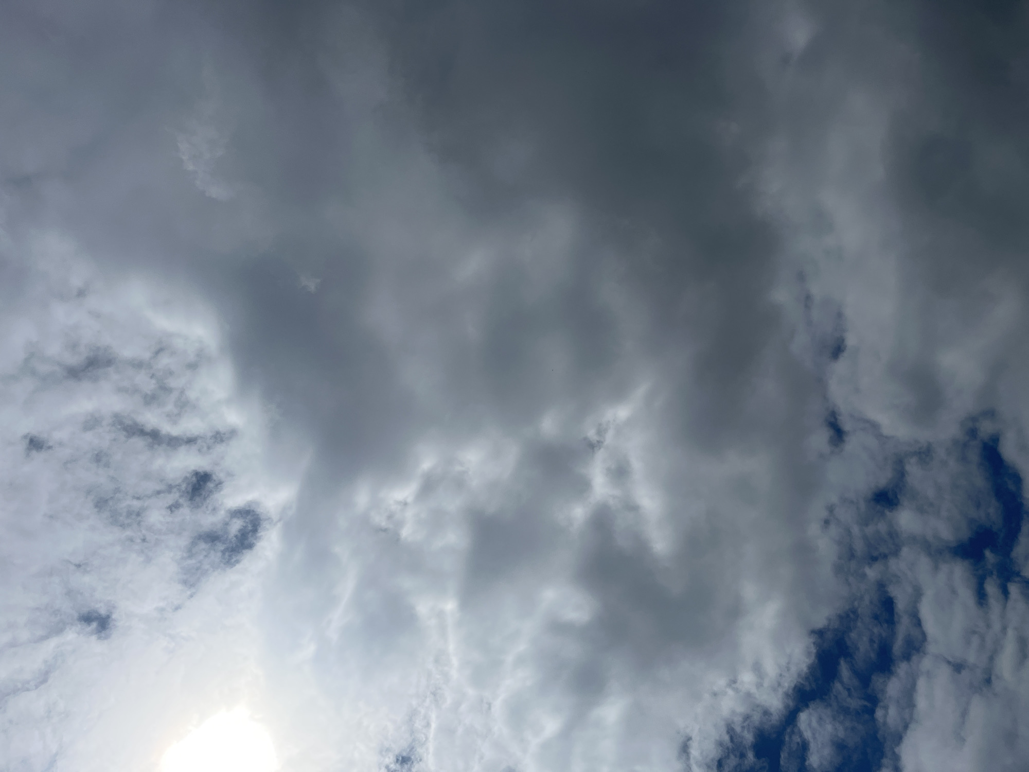 The image depicts a cloudy sky with varying shades of gray. Patches of blue sky are visible through gaps in the cloud cover. A bright light source, possibly the sun, is visible in the lower left corner of the frame. Location: Unknown, Unknown.