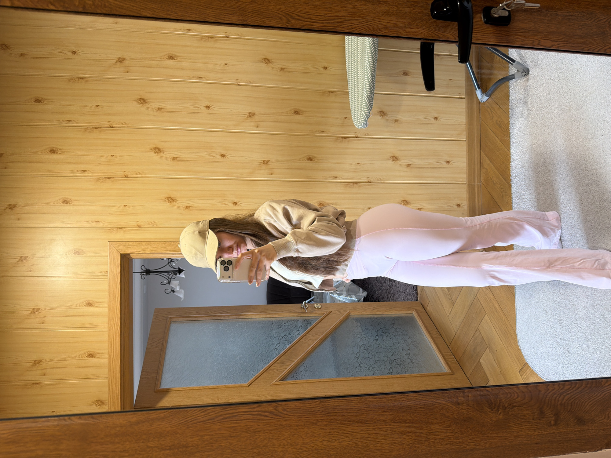 A woman is taking a selfie in a mirror. She is wearing a baseball cap, a beige sweatshirt, and pink pants. She is standing in a room with wood-paneled walls and a wood-framed doorway. The doorway leads to another room with a frosted glass door and a chandelier. In the mirror's reflection, there is a desk with keys and a lamp, as well as a carpeted floor. It is not possible to confirm the location in Buhovo, Bulgaria, based on visual information.