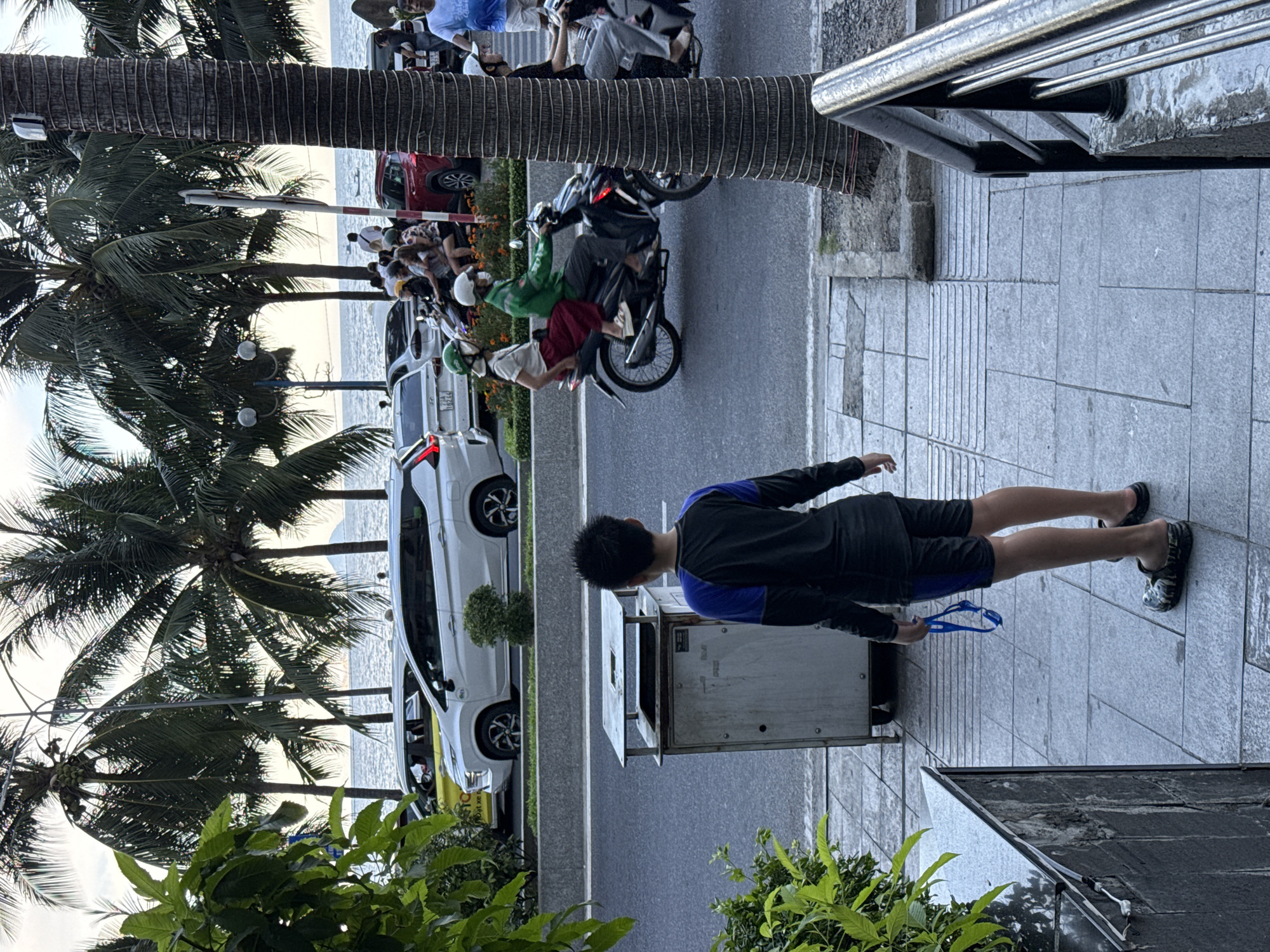 This image captures a street scene in Nha Trang Ward, Vietnam, with the ocean and palm trees visible in the background. In the foreground, a young boy stands facing away from the camera, holding a pair of swimming goggles. He is wearing a dark long-sleeved shirt with blue accents and dark shorts, and is barefoot except for a pair of patterned clogs. To his left is a metal trash receptacle. A paved sidewalk separates the boy from the street, where several vehicles, including cars and motorcycles, are present. On the motorcycles, people are seen wearing helmets and some are wearing masks. The atmosphere suggests a warm, possibly humid, day in a coastal city.
