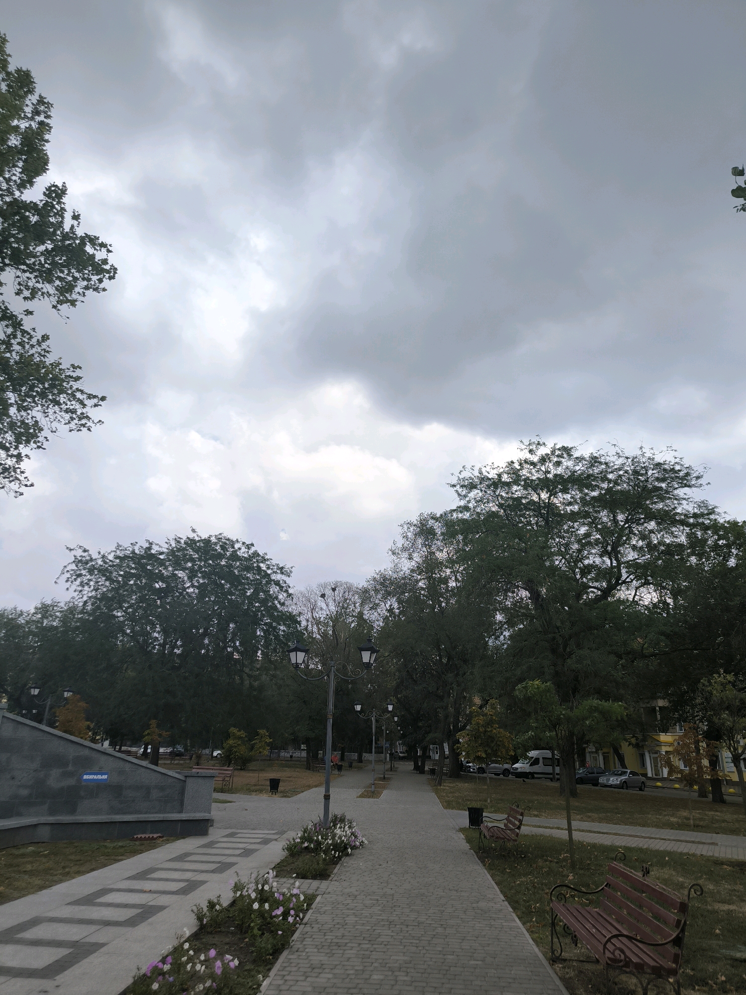 The image depicts a city park in Mykolaiv, Ukraine, under an overcast sky. A paved walkway runs through the park, flanked by trees and grass. Two empty wooden benches with ornate metal legs are visible on the right side of the walkway. In the background, there are more trees and some buildings. On the left side of the walkway, there is a gray stone monument with a blue plaque on it, and flowerbeds with pink and white flowers are planted along the path. Streetlights are also visible along the walkway. The scene appears to be quiet, with no people actively engaged in activities, though a few are faintly visible walking in the distance on the right. The overall impression is one of a serene, slightly melancholic atmosphere due to the cloudy weather.