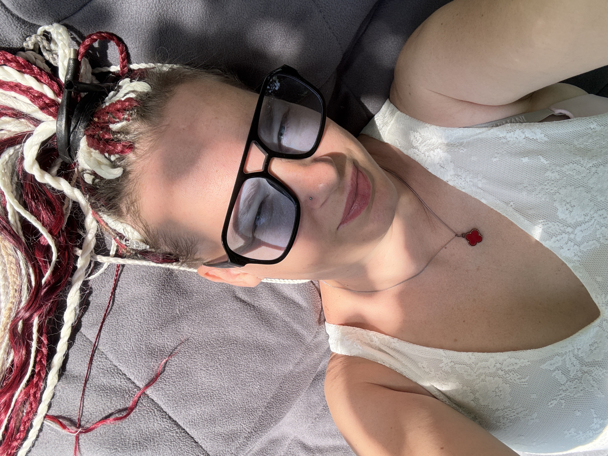 A young adult female is depicted in a close-up, overhead image, lying on her back on a quilted gray surface. Her gaze is directed towards the camera, accompanied by a slight smile. She wears prominent black-framed glasses with blue-tinted lenses, which reflect bright sunlight and partially obscure her eyes. A small silver stud is visible in her left nostril. Her lips are adorned with a muted pink color. Her hair is styled in synthetic braids of burgundy and off-white, secured in a bun on top of her head with a black band. She is dressed in a white, V-neck, lace-textured sleeveless top, revealing a thin silver-toned necklace featuring a small, red four-leaf clover pendant. Strong direct sunlight illuminates the scene, creating distinct highlights and shadows. The depicted setting is an unspecified interior or outdoor location within the city of Moscow, Russia.