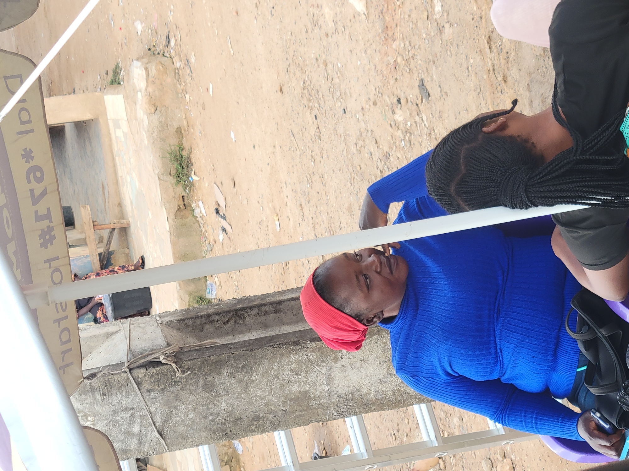 The image depicts an outdoor scene in Jos, Nigeria, under a white awning. A woman wearing a red headscarf and a blue turtleneck sweater is seated in a purple chair. She is looking to her right, with her hand near her face. To her right, another person with braided hair is visible from behind, wearing a black t-shirt. A black bag rests on the chair next to the woman in blue. In the background, a concrete pillar is visible, along with a ladder. The ground is sandy and littered with debris. An inscription is partially visible on the awning.