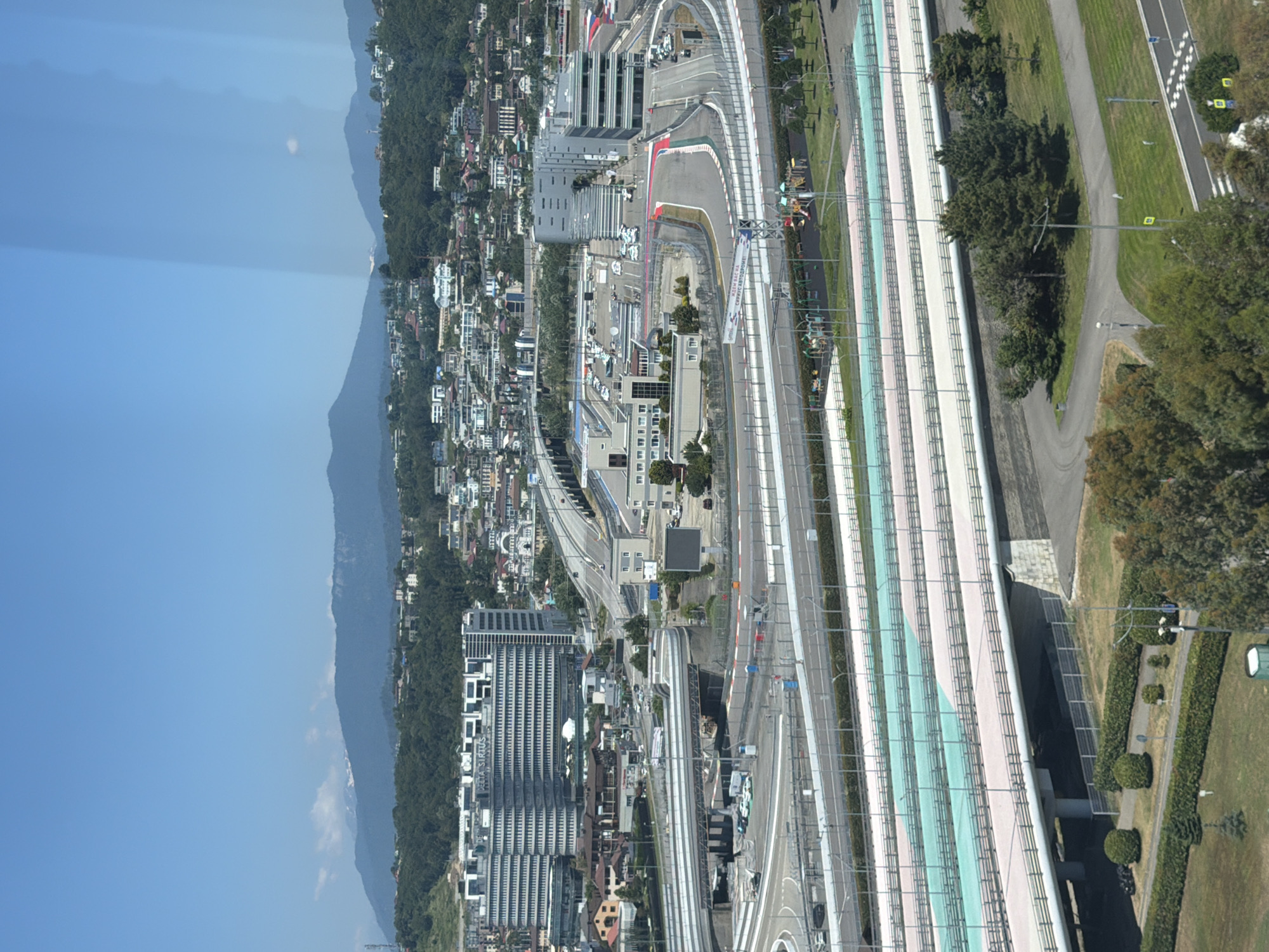 This is an aerial, rotated, landscape photograph of a modern race track, likely a Formula 1 circuit, situated in a coastal mountain region. The track features various colored racing lanes and prominent white lines marking its boundaries. Adjacent to the track are several multi-story buildings, some with modern glass facades and others with more traditional architecture, nestled among dense green trees and hillsides. A clear blue sky dominates the upper left portion of the image, suggesting it's daytime with fair weather. The overall scene appears to be a well-developed urban or resort area, characterized by sleek infrastructure and natural mountainous terrain.
There is some visible text on a banner on the side of the track. The most prominent part of the text reads "СПРАВЕДЛИВАЯ АВТО..." which translates to "FAIR AUTO..."
The location is contextualized as Sirius, Russia.