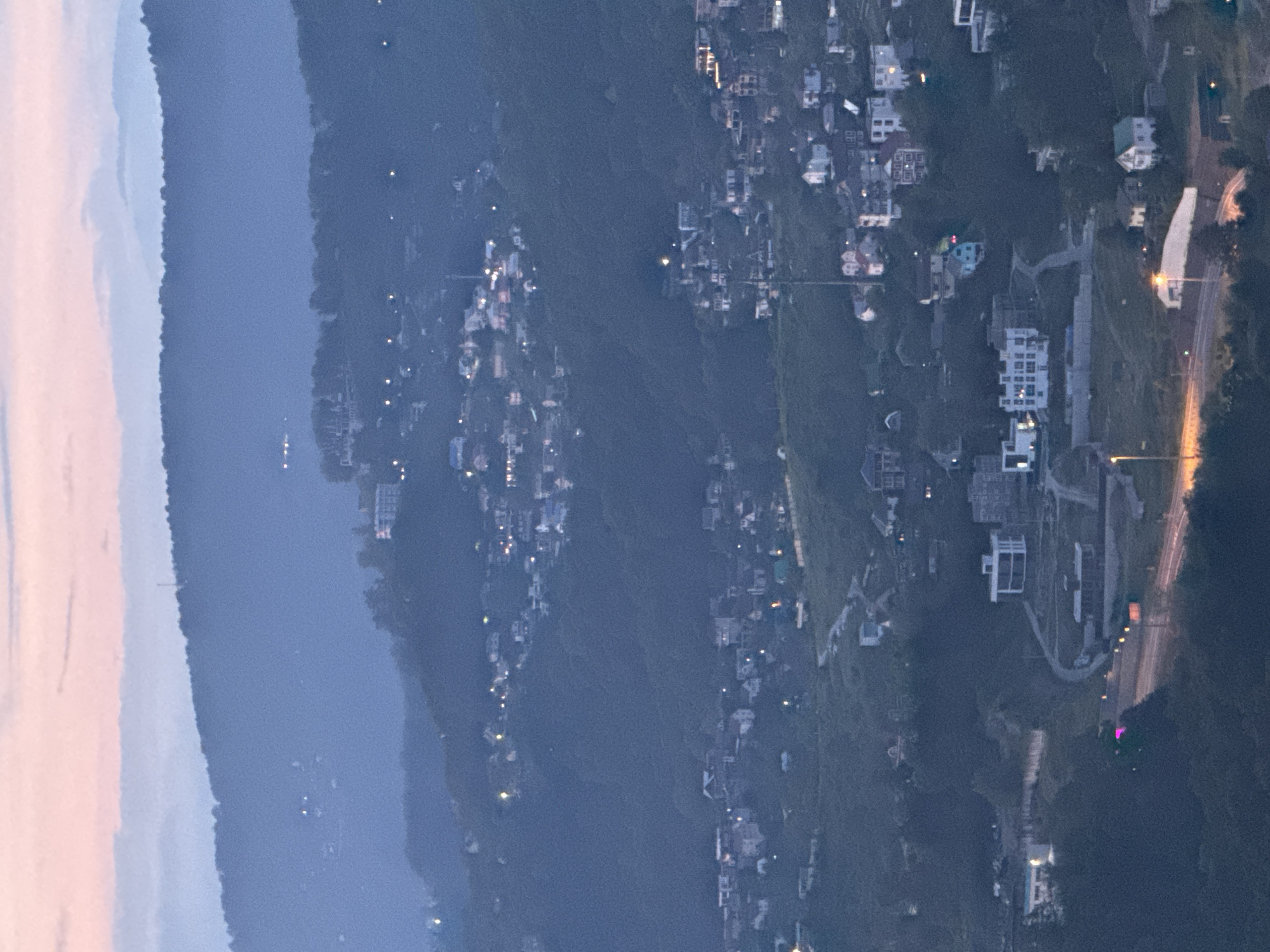 This image depicts a twilight aerial view of a hillside settlement in Sochi, Russia. The sky displays a gradient of soft pink and pale blue hues, indicative of sunset or sunrise. Numerous houses and buildings are scattered across the undulating terrain, many illuminated with warm, interior lights. A road network winds through the settlement, with streetlights visible along some stretches. The distant landscape is characterized by layered, mist-shrouded mountains, creating a sense of depth. The overall atmosphere is serene and captures the transition from day to night in a residential area nestled within a natural landscape.