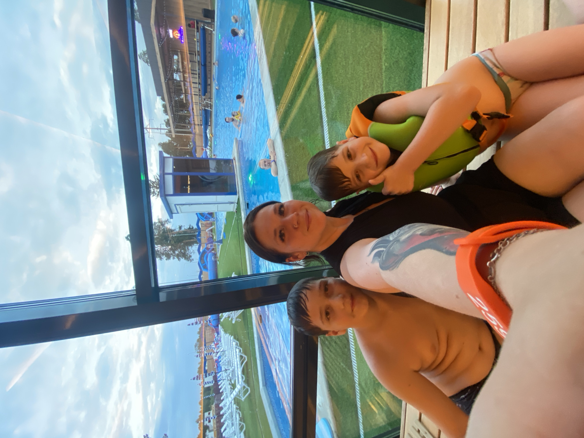 A woman and two young boys are posing for a selfie in front of a large window overlooking an outdoor swimming pool complex. The woman is in the center, wearing a black dress, and has tattoos on her arm. The boy on the left is shirtless, and the boy on the right is wearing a green and orange life vest. The swimming pool complex features lounge chairs, umbrellas, and blue inflatable water slides. In the background, several people are visible swimming in the pool. The sky is cloudy. This scene is likely at a public water park or resort in the city of Votkinsk, Russia, given the context.