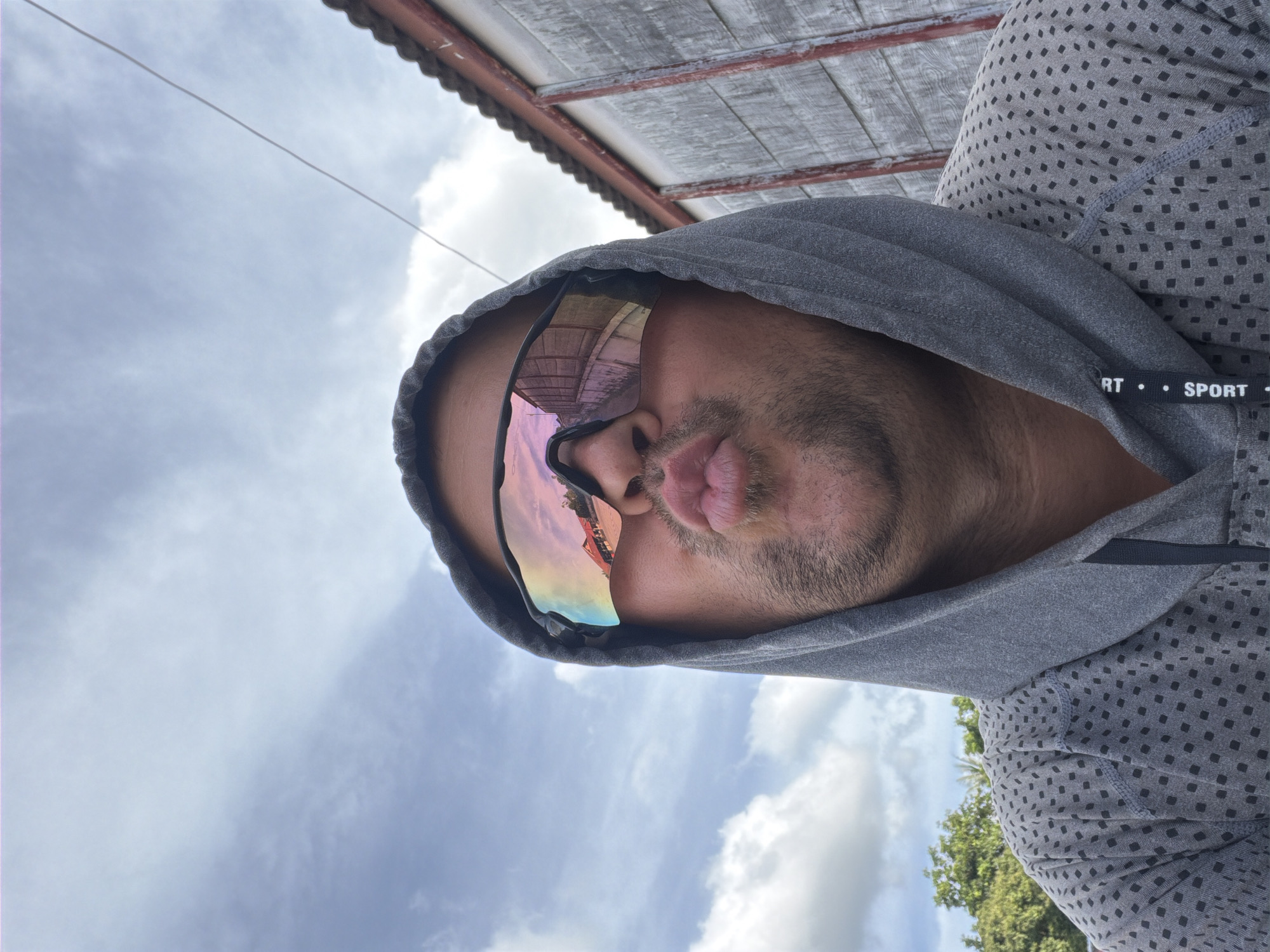 A man, wearing a grey hooded sweatshirt and mirrored sports sunglasses, puckers his lips for a selfie. The sunglasses reflect a building with a red roof and a fence. He is outdoors with a cloudy sky above and greenery and a weathered wooden structure in the background. The location is identified as Baan Mae Nam, Thailand.