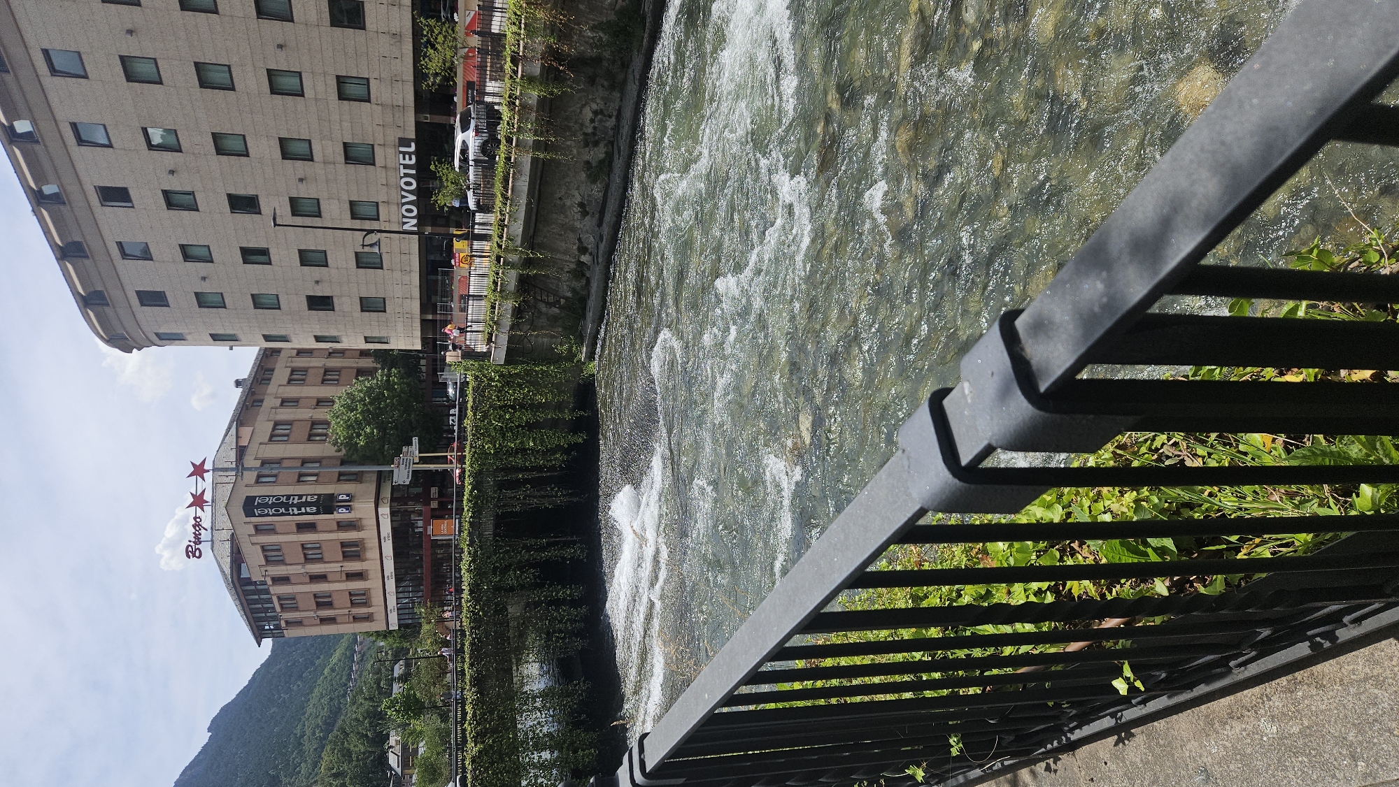 This detailed image depicts a daytime scene along the Gran Valira river in Andorra la Vella, Andorra. In the immediate foreground, a black metal railing with vertical bars and a horizontal top bar extends from the left, positioned above green ground cover and a concrete path.
Beyond the railing, the Gran Valira river flows rapidly from left to right, exhibiting clear water with visible rocks on its bed and several areas of white rapids and turbulence. In the mid-ground, a bridge spans the river, heavily draped with thick green climbing vines that hang downwards towards the water. Above the bridge, a paved street runs parallel to the river, featuring a black metal barrier on its edge, several indistinct pedestrian figures, and parked vehicles, including a white SUV on the right.
The background is dominated by urban structures and natural topography. To the left, a multi-story, light brown building with numerous windows displays a "Bingo" sign topped by two red stars on its roof, and a vertical banner on its facade reading "arthotel" with two "P" symbols below. To the right, a taller, lighter-colored multi-story building with a regular pattern of rectangular windows prominently features a "NOVOTEL" sign on its ground level. In the far background, a steep, densely forested green mountain slope rises on the left side under a bright sky with scattered white clouds.