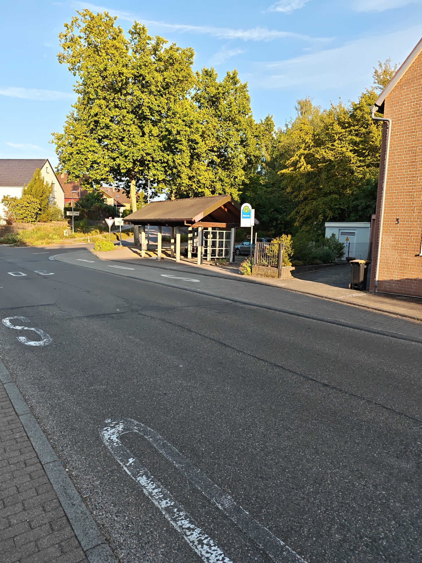 The image depicts a street scene in Nordheim, Germany, featuring a bus stop. The bus stop shelter has a wooden roof supported by posts and contains seating. A blue and white bus stop sign with an "H" symbol is visible. The street is asphalt, with white markings indicating lanes and pedestrian crossings. On the right side of the street, a brick building with a visible house number "3" is present, along with a trash bin. In the background, there are trees with green foliage and several houses. The sky is blue with scattered clouds. The overall environment suggests a residential area during daytime.
