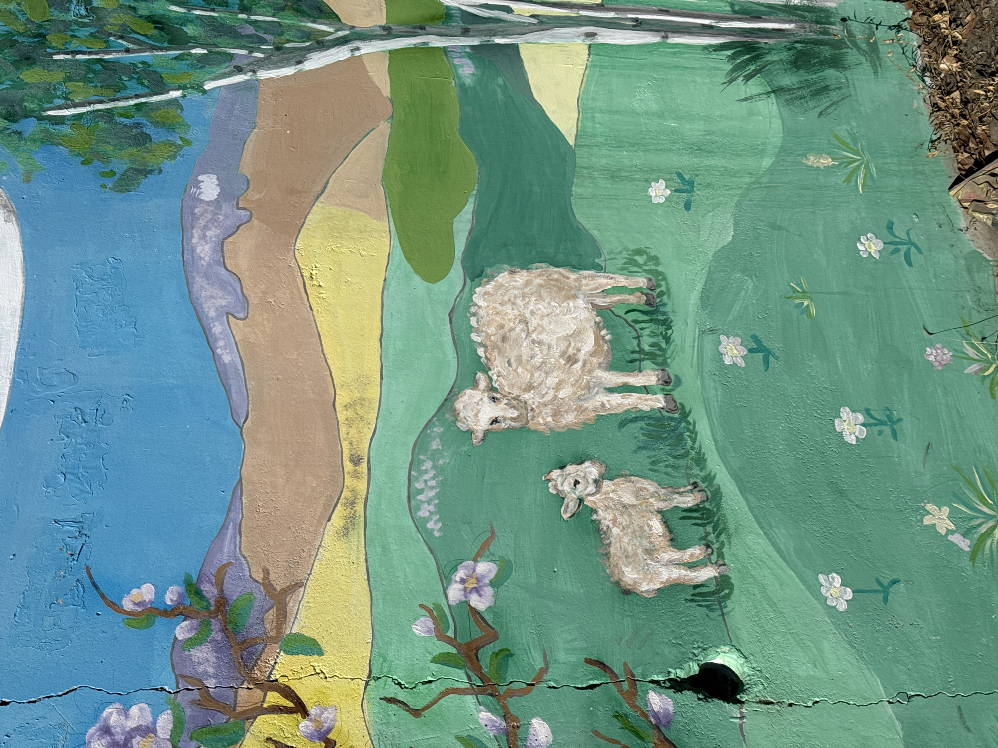 The image shows a section of a painted mural on a concrete surface. The mural depicts a pastoral scene with a blue sky on the left, a large tree trunk with brown, yellow, and green sections, and a green meadow on the right. In the meadow, there are two sheep lying down, one larger than the other. White flowers are scattered throughout the meadow. There are flowering branches with purple blossoms in the foreground. The concrete surface has cracks running across it. The mural is situated in Borzya, Russia.