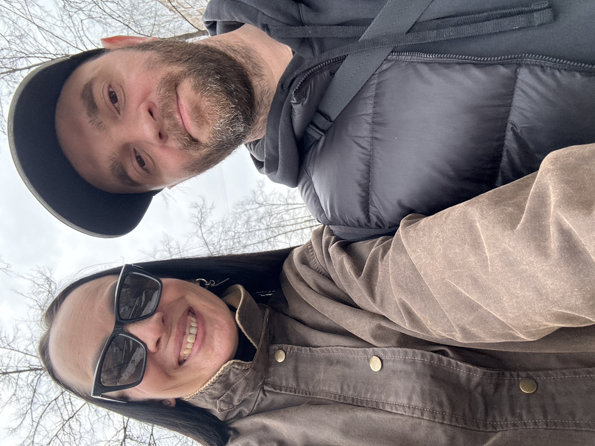 A close-up selfie captured in Chaikovsky, Russia, features two adults: a female on the left and a male on the right. The female has long dark hair, wears black cat-eye sunglasses, a brown, high-collared jacket with brass-colored snap buttons, and a dark garment underneath. She is smiling, revealing her teeth, and has a silver-colored earring on her left ear. The male has a beard with visible graying, wears a dark baseball cap, a black puffer vest over a black hooded sweatshirt, and a dark shoulder strap across his chest. His expression is neutral. Both individuals are looking directly towards the camera. The background consists of bare tree branches against an overcast sky, with visible patches of snow on the ground and faint building structures in the distance, indicating an outdoor, urban setting in a cold season.