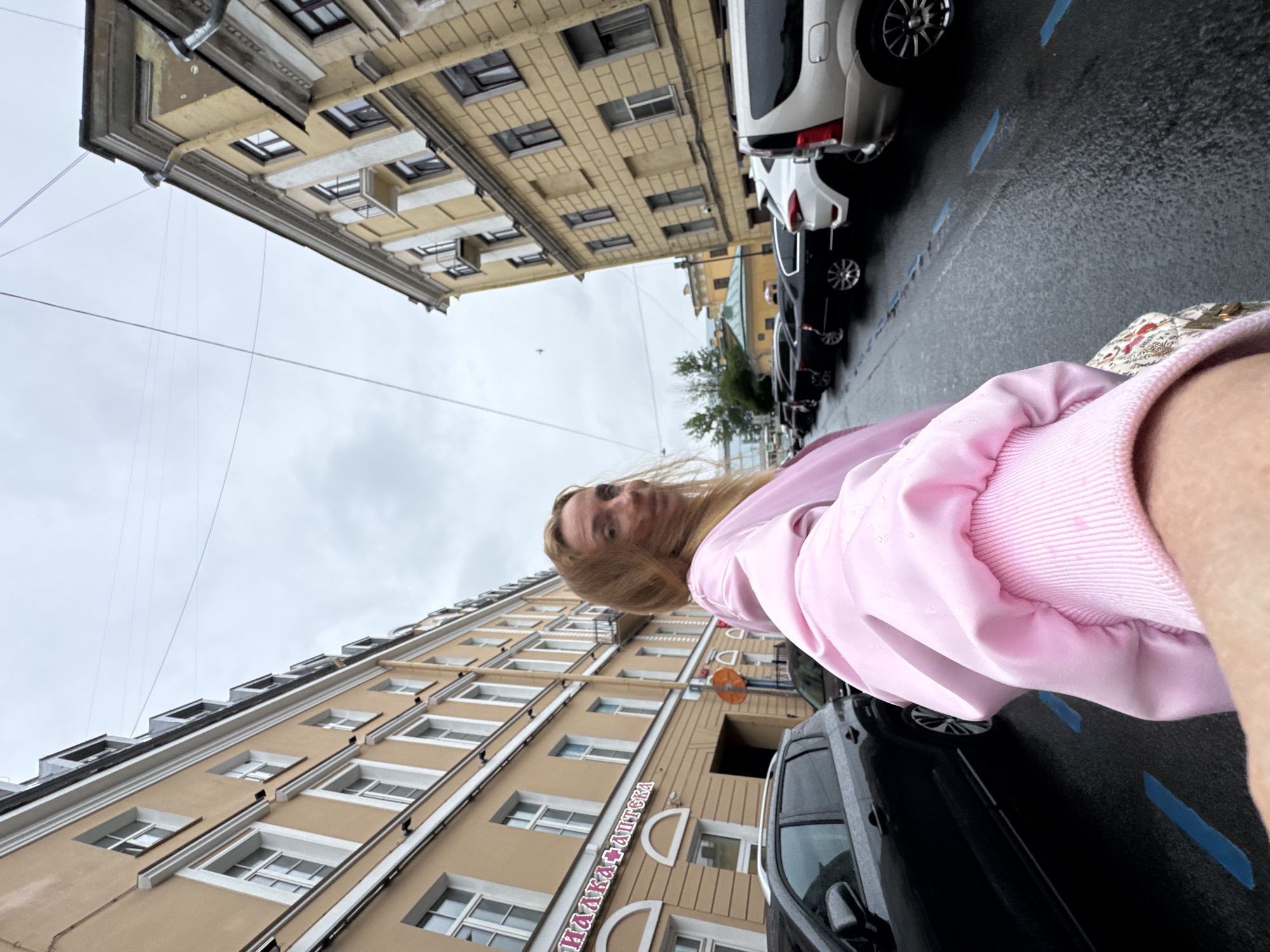 A woman is taking a selfie in a city street in Saint Petersburg, Russia, on a cloudy day. The woman is wearing a pink jacket. She is standing on the right side of the frame, with her right arm extended towards the camera. The street is lined with multi-story buildings on both sides. On the left side of the street, there is a building with a sign that says "АПТЕКА". The street is wet, indicating that it has recently rained or is raining. There are several cars parked along the street. The sky is overcast, and there are power lines visible overhead.