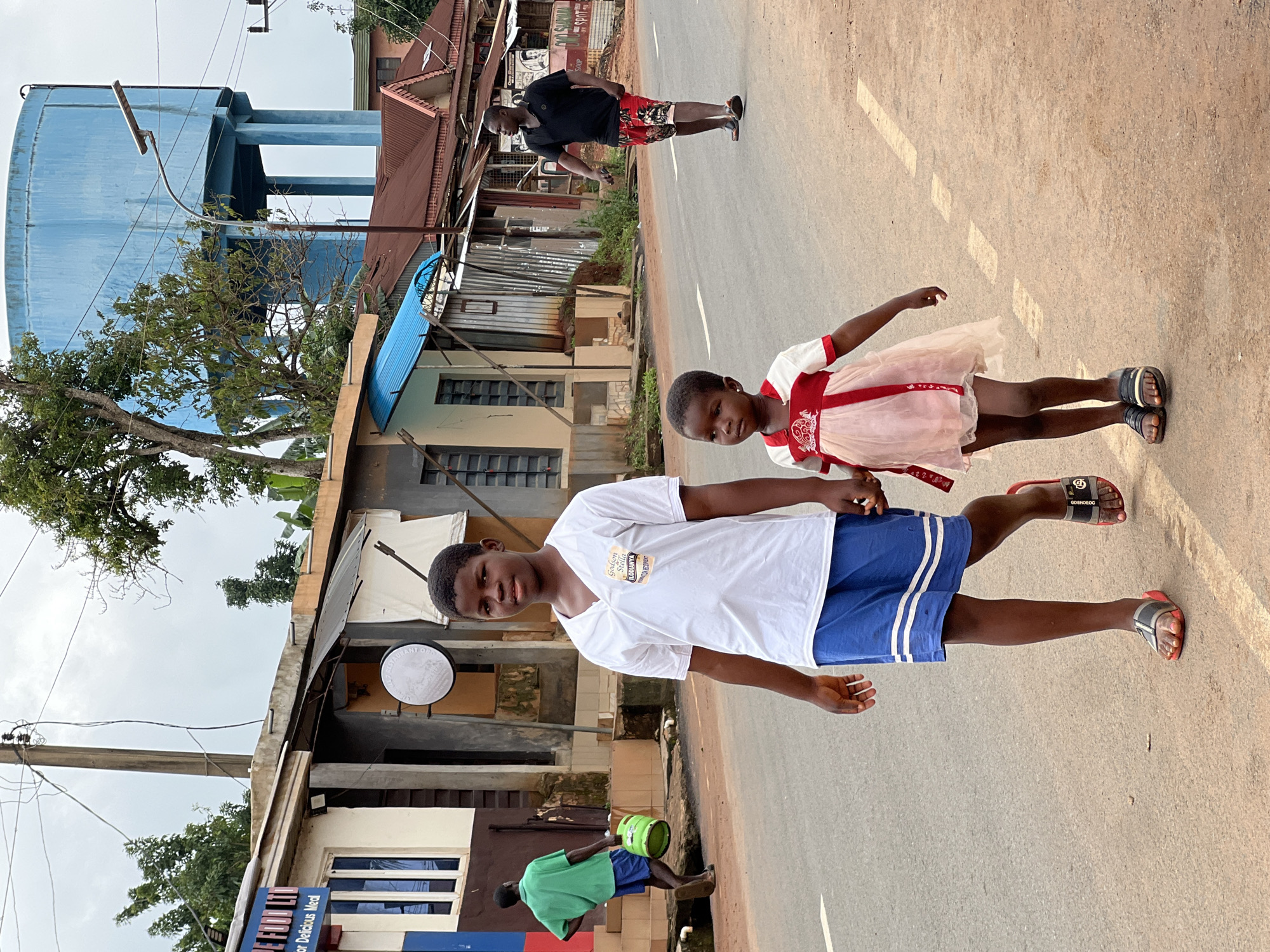 In Nkpor, Nigeria, two children, a boy and a younger girl, walk hand-in-hand on the right side of a paved road. The boy, wearing a white t-shirt and blue shorts, is positioned in the foreground and faces the camera. The girl, in a pink and white dress, is to his right and slightly behind him. Behind them, on the left side of the road, a man in a green shirt and blue shorts carries a green bucket. Further down the road on the right, a man in a black t-shirt and patterned shorts walks away from the camera, looking to his left. The background shows a row of buildings with various architectural styles, including a structure with a blue corrugated metal roof. A tall, blue water tower stands in the distance, visible above the buildings and trees. The sky is overcast.