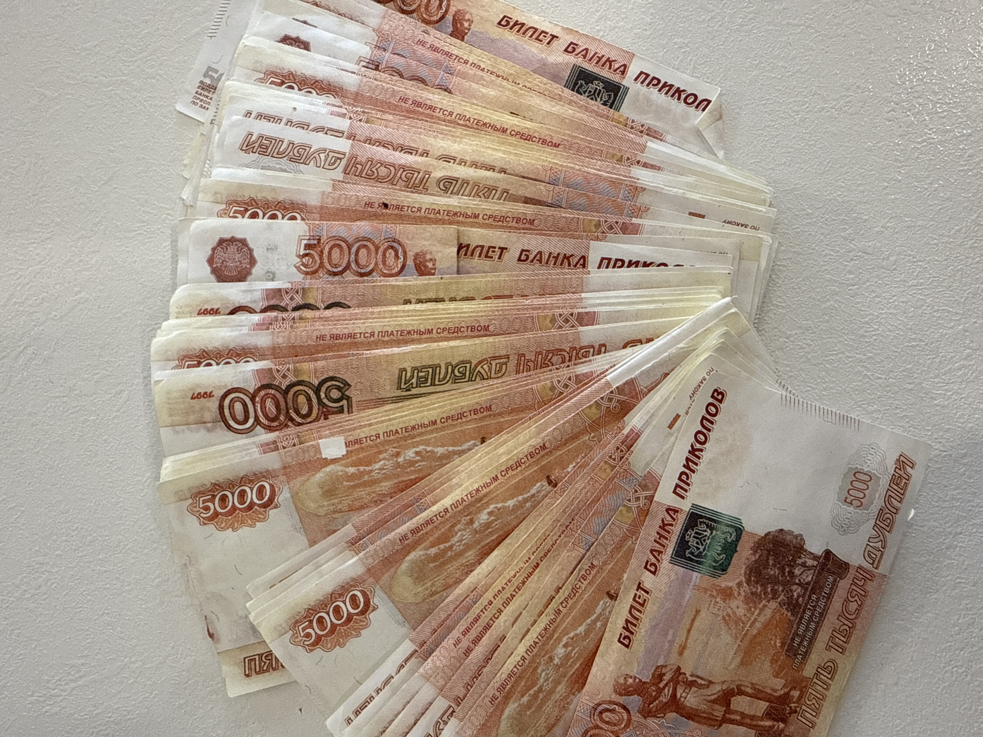 The image shows a fanned stack of "Bank of Prank Tickets" (БИЛЕТ БАНКА ПРИКОЛОВ) designed to resemble Russian 5000 Ruble banknotes. Each note is marked with "НЕ ЯВЛЯЕТСЯ ПЛАТЕЖНЫМ СРЕДСТВОМ," indicating they are not legal tender. The dominant color is reddish-orange, with the number "5000" prominently displayed. The phrase "ПЯТЬ ТЫСЯЧ ДУБЛЕЙ" (Five Thousand Doubles) is visible on the notes, further emphasizing their non-legal tender status. The bills are arranged in a disorderly fashion, with edges overlapping. The backdrop is a light-colored, textured surface. The image does not include any information about the location in the city of Votkinsk, Russia.