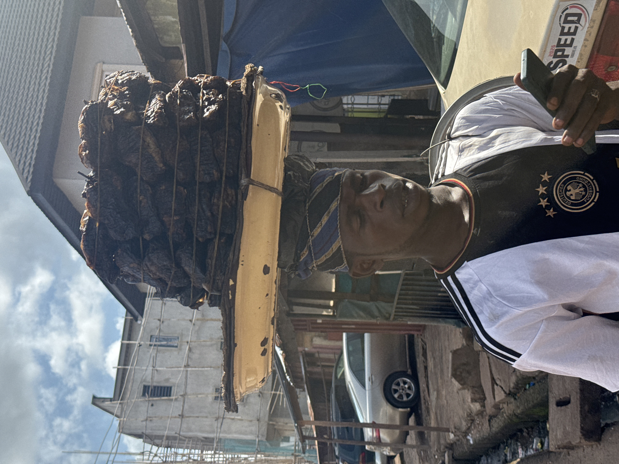 A man balances a large stack of dried fish on his head, with a metal tray underneath, in the city of Enugu, Nigeria. He is wearing a black and white t-shirt and a patterned headwrap. In his right hand, he holds a smartphone. Behind him, construction is underway on a building. A silver sedan is parked to his left, and a dusty, light-colored car is partially visible on the right. The ground appears to be unpaved and there is a drainage ditch visible. The sky is cloudy.