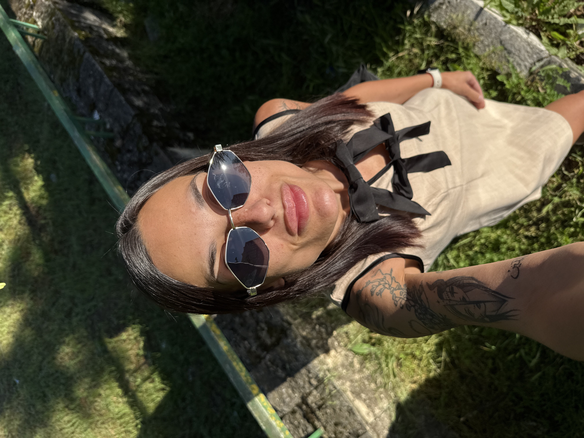 This media file captures an outdoor portrait of a woman relaxing on green grass in Pleven, Bulgaria, under bright sunlight. The image is taken from an angled perspective, showing the woman lying diagonally across the frame, looking towards the camera. She has long, dark hair and is wearing large, geometric-shaped sunglasses with dark lenses and thin metallic frames. Her lips are slightly parted, displaying a pinkish hue.
She is dressed in a light-colored, likely beige or off-white, dress with black straps and multiple black bows tied decoratively on the upper part. Her left arm and forearm are adorned with several dark, intricate tattoos, including a prominent design of a stylized face or character. A white smartwatch is visible on her left wrist.
The setting appears to be a park or garden, with lush green grass covering much of the ground. Visible in the background are a concrete or stone border and a green metal railing. The strong sunlight creates distinct shadows, indicating a clear, sunny day. The woman's expression is relaxed and content, suggesting she is enjoying the moment or posing casually for the camera. There is no visible text in the image.