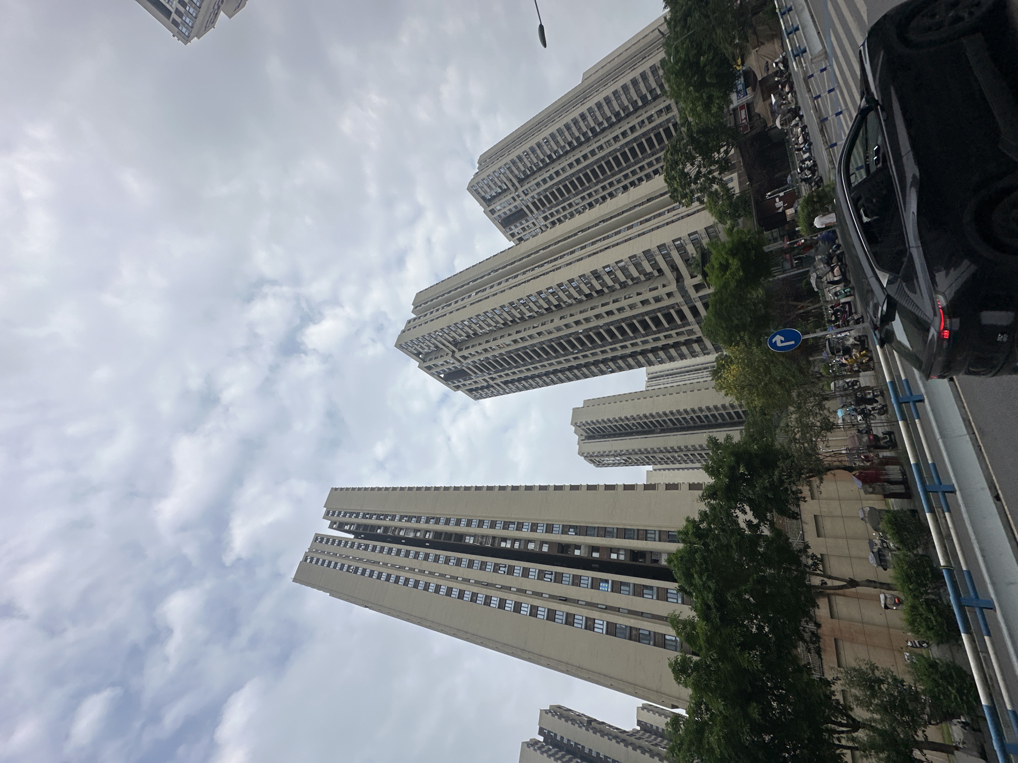 The image depicts a street scene in Chongqing High-tech Zone, China. Tall, modern apartment buildings dominate the skyline. A dark-colored car is partially visible on the right side of the frame. A street with trees lining the side and parked motorcycles are seen. The sky is overcast with cloud cover. A street sign is observed along the road.