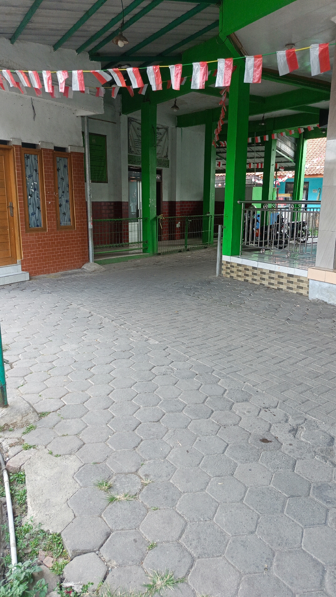 The image depicts an outdoor scene in front of a building with a green roof and pillars. The foreground is dominated by a hexagonal paver walkway. In the middle ground, there is a building with red brick walls and windows, and a green metal fence. Red and white flags are strung across the top of the building, suggesting a celebration or national holiday. In the background, there are more flags, a motorcycle parked under a sheltered area, and a glimpse of a tiled roof. The overall impression is of a public space, possibly a place of worship or a community center. The lighting suggests it is daytime.