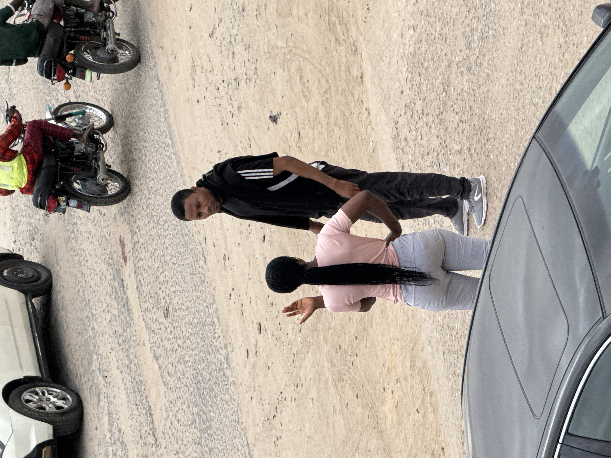 The image shows a street scene in Lugbe, Nigeria. A man and a woman are standing on a sandy roadside next to a dark gray sedan. The man is wearing a black Adidas track jacket and gray athletic shoes. The woman is wearing a pink t-shirt and gray leggings. Her hair is styled in a long braid. The woman has her hand up and is facing the sedan. Two motorcycles are parked on the roadside. A white vehicle is also present in the image.