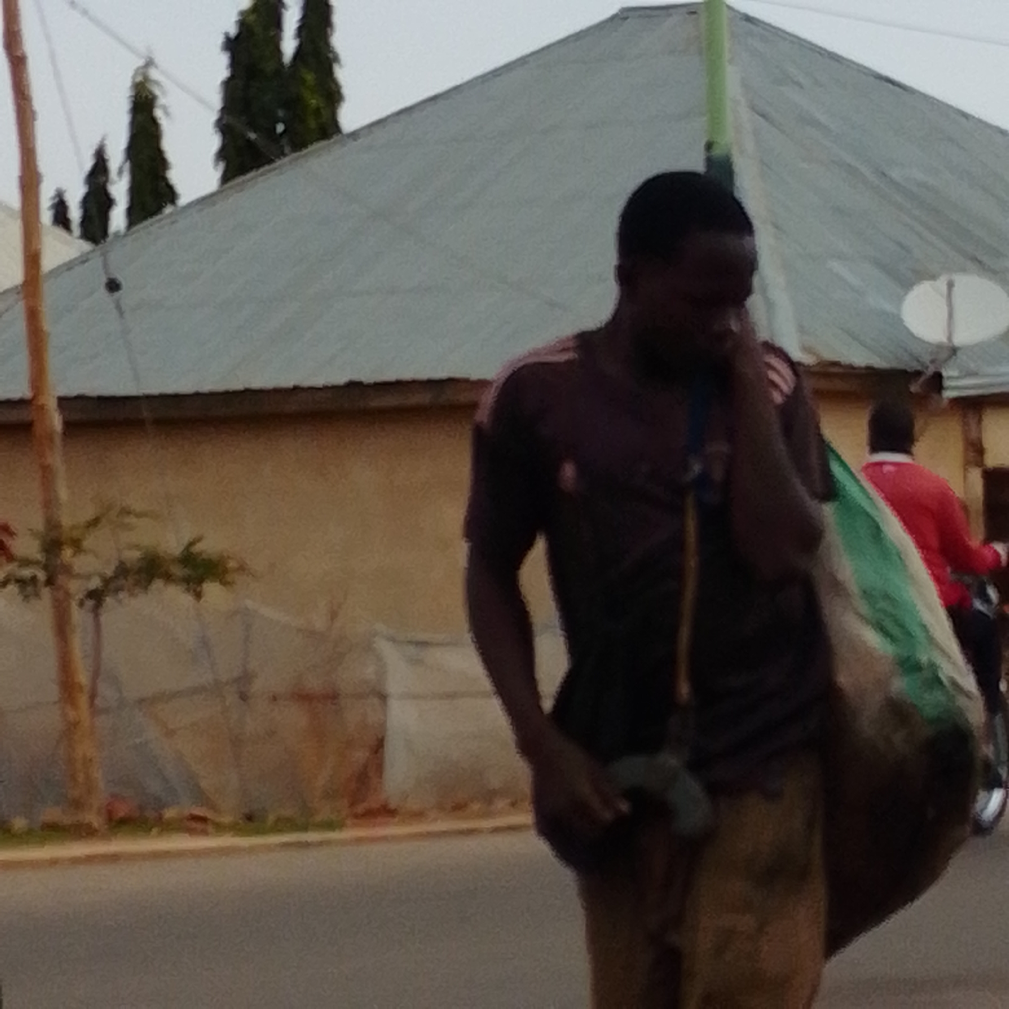 The image shows a person walking on a street, carrying a large bag over their shoulder. The person is wearing a dark t-shirt and tan pants. The bag appears to be made of a green and tan material and is quite large. In the background, there is a building with a gray, sloped roof, and some trees. There is also a second person in the background wearing a red shirt, and what appears to be a motorcycle. A satellite dish is visible on the roof of the building. The lighting suggests it is daytime, possibly overcast. The scene appears to be in an urban or semi-urban environment. No visible text is present in the image. The overall impression is of someone going about their daily business, likely transporting goods or belongings. The location context suggests this scene is in Gombe, Nigeria.