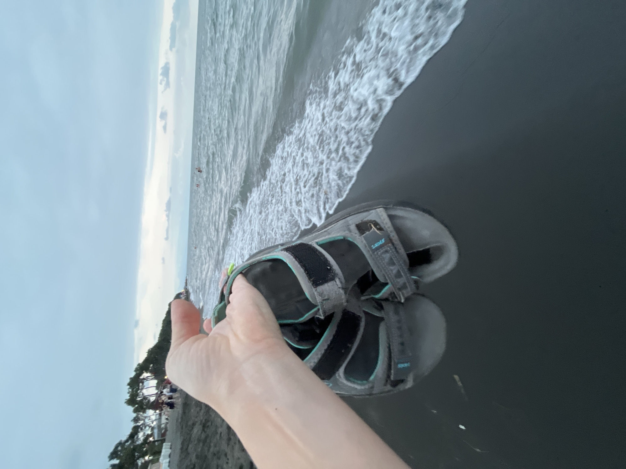 A hand holds a pair of gray and teal sandals over a black sand beach. The ocean waves are rolling in, and the sky is overcast. In the background, trees line the shore, and a few people are visible in the water. The scene appears to be from Ureki, Georgia, a resort town known for its magnetic black sand beaches.