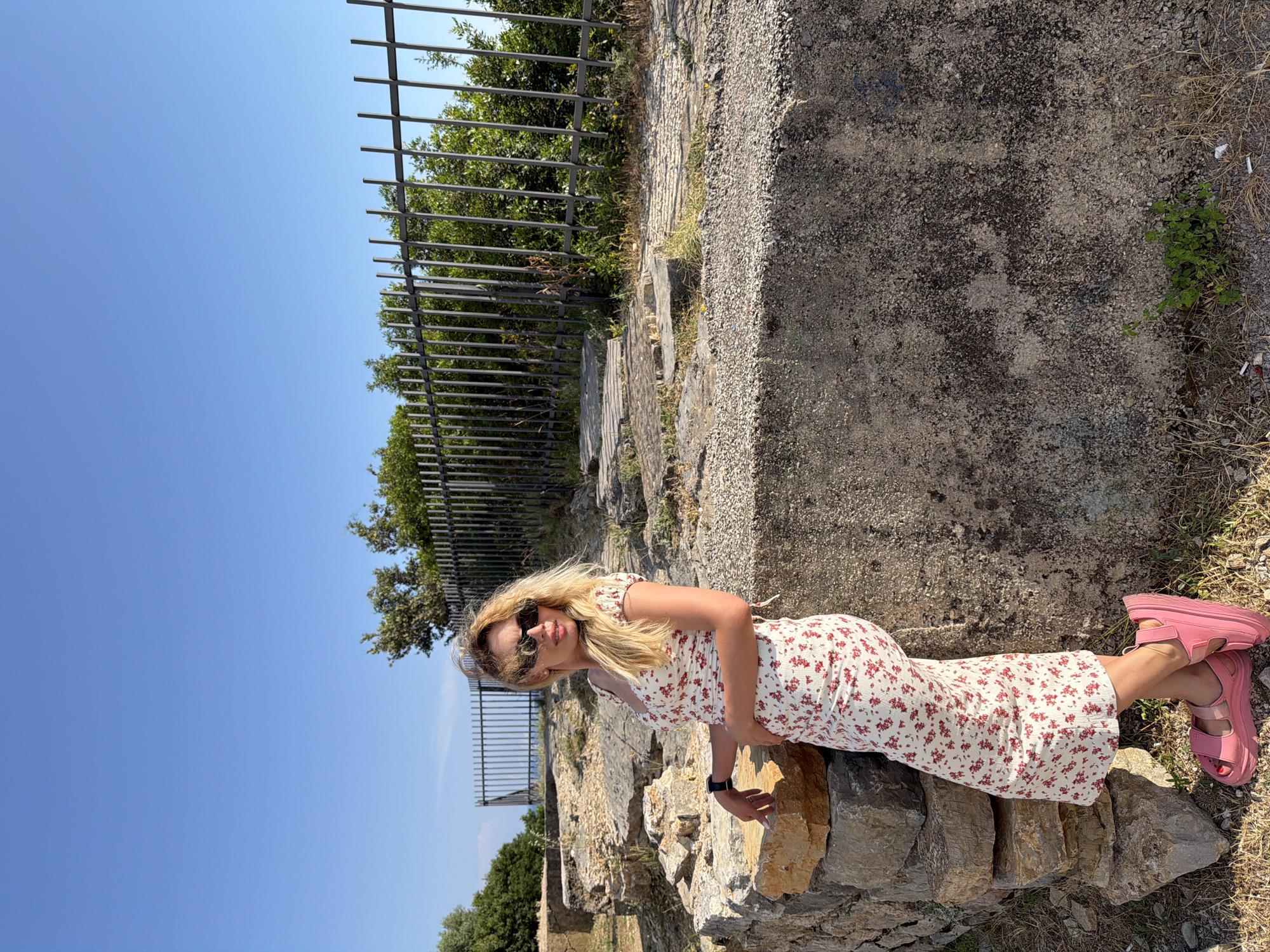 An outdoor, eye-level, full-shot photograph captures a woman standing in the ruins of an ancient site. She is positioned to the left of the frame, leaning against a rough stone wall. The woman has fair skin, blonde hair styled in a high ponytail, and wears large sunglasses. Her attire consists of a white, fitted midi dress adorned with a red floral pattern, and bright pink chunky platform sandals. She is looking off to her right, with her left hand resting on the wall and her right hand on her hip.
The environment appears to be an archaeological site under a clear blue sky. In the foreground, there are weathered stone structures and loose rocks. Behind the woman, a tall, dark metal fence stretches horizontally across the frame, with lush green trees and foliage visible beyond it. The ground is covered in dry grass, gravel, and scattered stones, suggesting a natural and somewhat unkempt setting. The overall impression is one of a historical location bathed in bright sunlight. The location is identified as being in Budva, Montenegro.