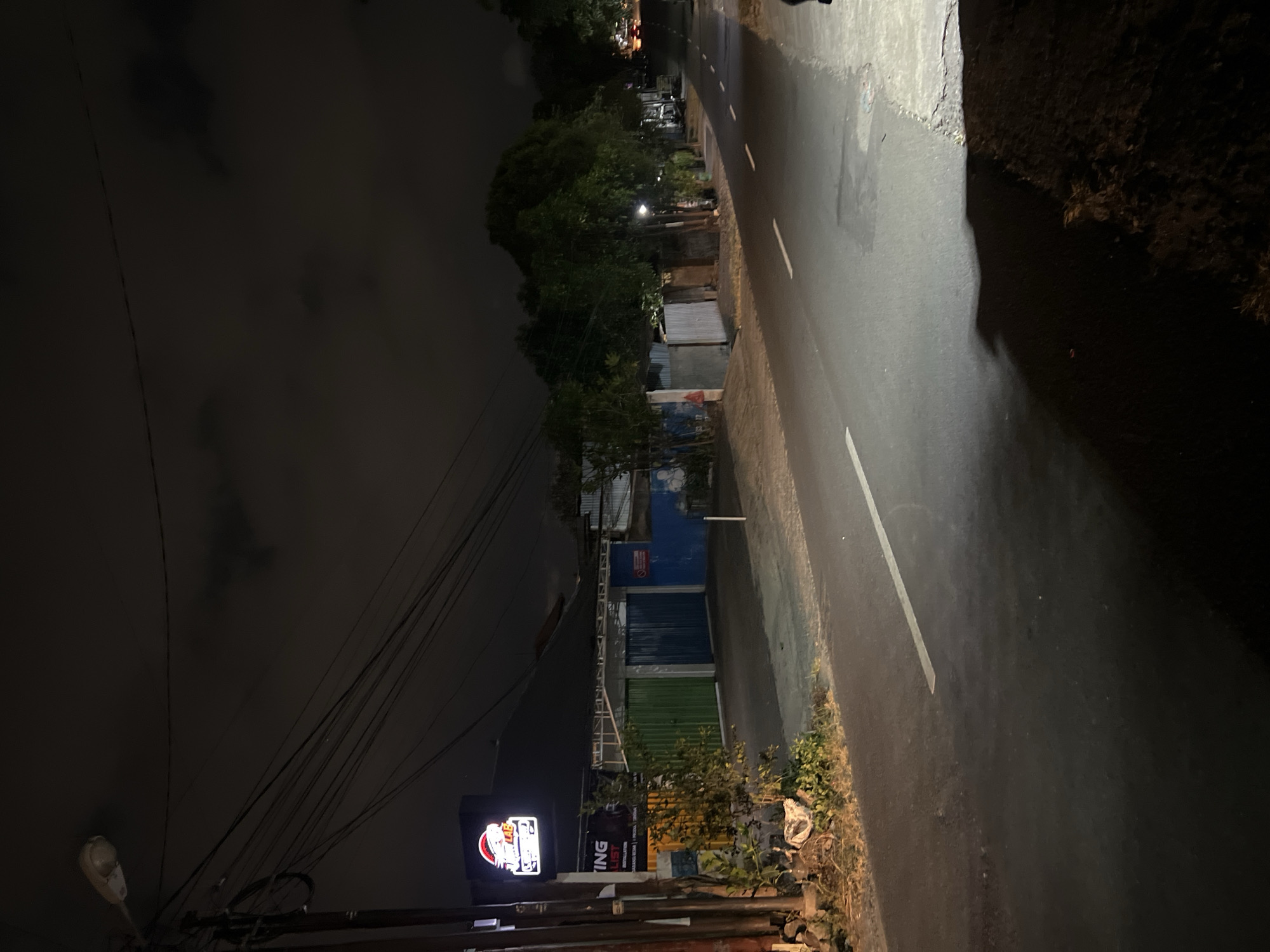 The image depicts a street scene at night in Denpasar, Indonesia. The foreground shows a dark asphalt road with a white painted line dividing the lanes. On the left side of the road, there is a dirt shoulder with some dry vegetation. A prominent pole with electrical wires and a streetlamp is visible on the far left. Behind the shoulder, there is a building with a dark roof and a brightly lit sign that reads "KLUB" and "LAB" with a stylized image. Further back, the building has doors and windows, some of which appear to be colored in green, blue, and white. The background features trees and other structures, with some distant lights indicating further development. The sky is dark with some cloudy formations. The overall scene suggests a typical urban or semi-urban environment at night, possibly a commercial area given the illuminated signage.