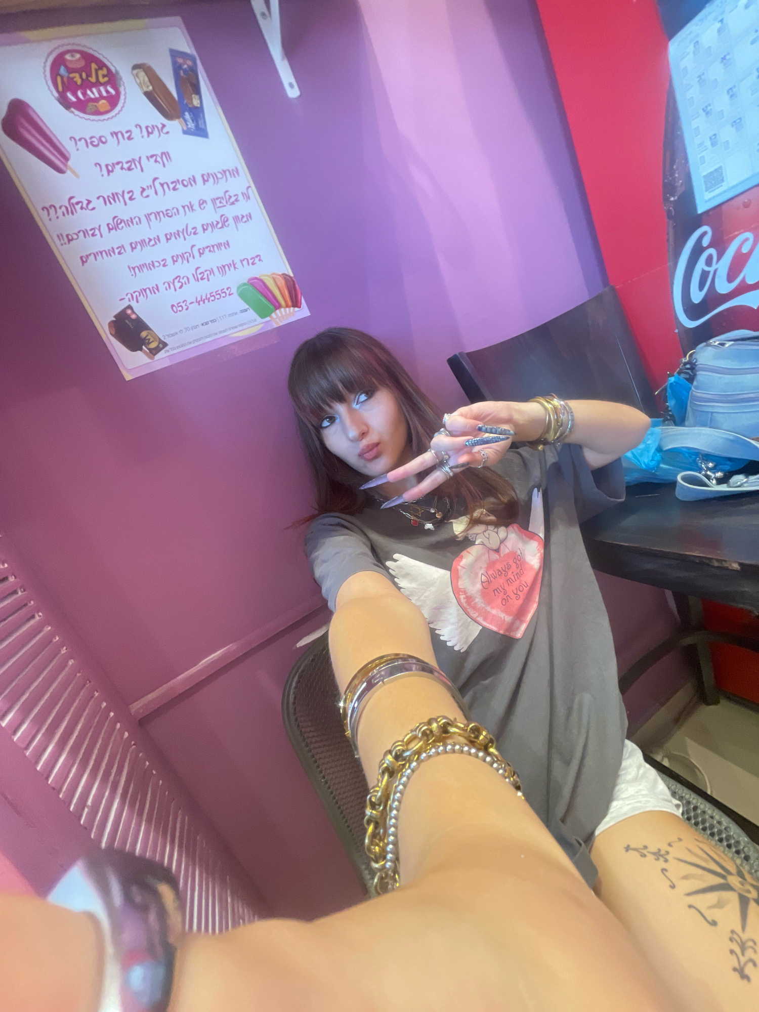The image shows a young woman with long dark hair and bangs, wearing a gray t-shirt with a graphic of a winged heart that says "Always got my mind on you." She has long, pointed nails with rhinestones. She is wearing multiple bracelets on her left wrist and rings on her fingers. She is holding up two fingers in a peace sign. The background is a pink wall with a poster advertising ice cream with Hebrew text. To the right, a portion of a Coca-Cola branded refrigerator is visible. The woman is sitting in a chair at a table. A blue bag and other items are on the table. The image is taken from a low angle, looking up at the woman. The scene appears to be indoors, possibly in a shop or cafe. The poster provides an indication that the location is in Raanana, Israel, as the text is in Hebrew and includes an Israeli phone number.