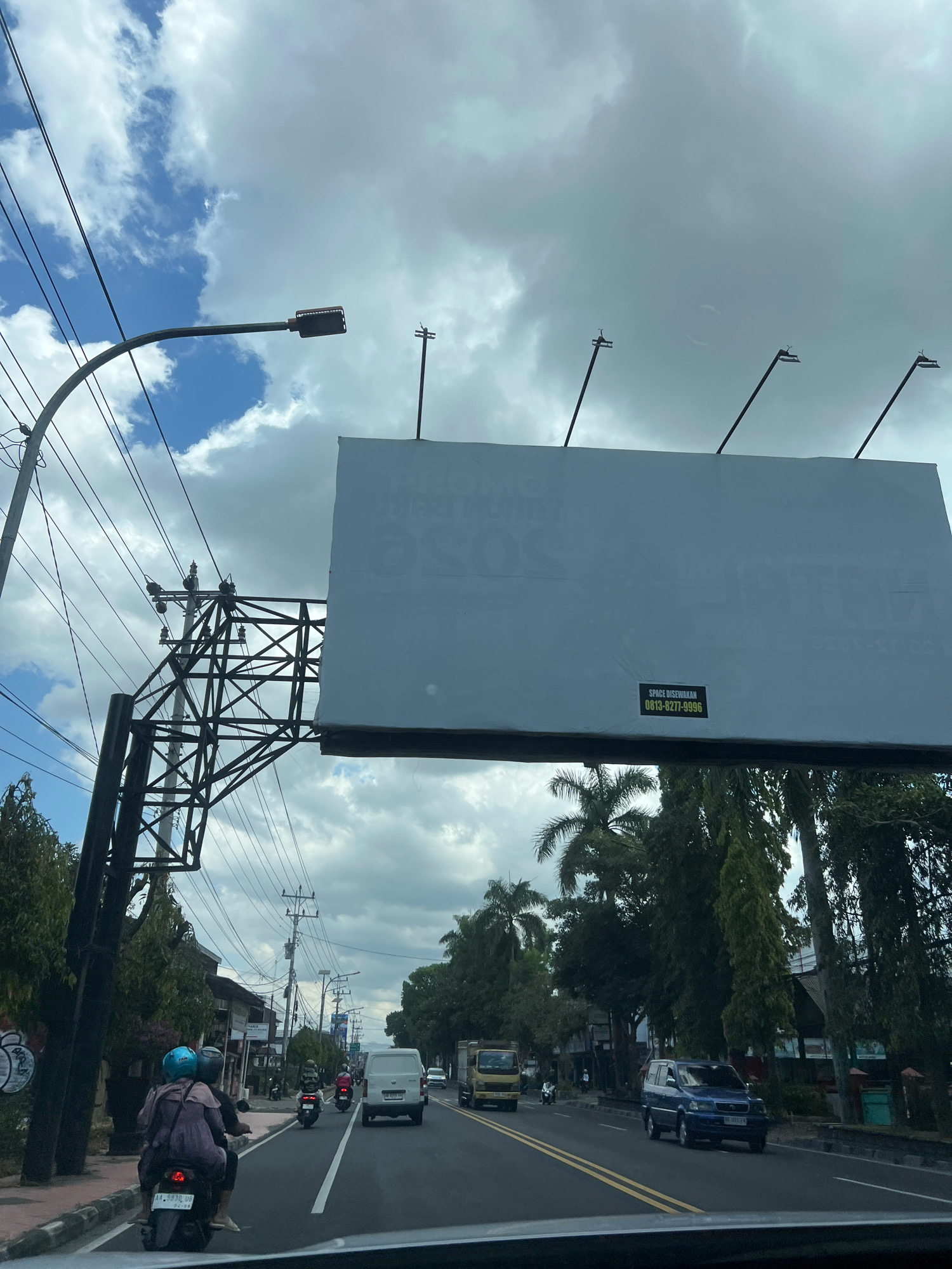This is a vertical, eye-level shot taken from a moving vehicle, likely a car, on a road in Magelang, Indonesia. The sky is filled with dramatic, heavy grey clouds, with patches of blue sky peeking through. A large, blank white billboard dominates the upper half of the frame, supported by a sturdy black metal structure. The billboard has a small black and yellow sticker on its lower right corner with white text that reads "SPACE DISEWAKAN 0813-8277-9996". On the left side of the image, a curved lamppost with a street light extends over the road, and a complex network of black electrical wires is visible, strung between utility poles.
Below the billboard, the scene depicts a busy street. Several motorcycles are present, including one in the foreground carrying two individuals wearing helmets. A white van drives in the right lane, followed by a blue SUV. Other vehicles and motorcycles are visible further down the road. Trees line the sides of the street, with some palm trees among them. Buildings can be seen in the background, suggesting a commercial or residential area. The road itself has clear lane markings. The overall impression is of a typical day in a moderately busy Indonesian city.