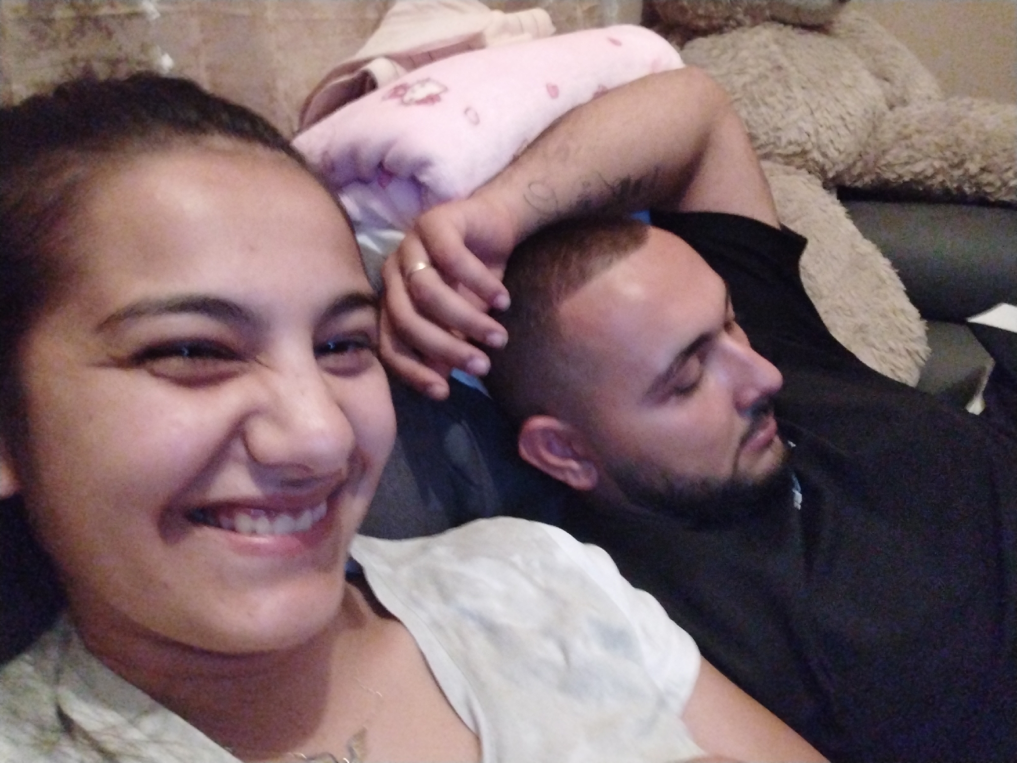 The image shows two people lying on a couch. The person on the left is smiling, and the person on the right appears to be sleeping. There's a teddy bear and a pink blanket on the couch near the sleeping person. The location in the city is unknown.