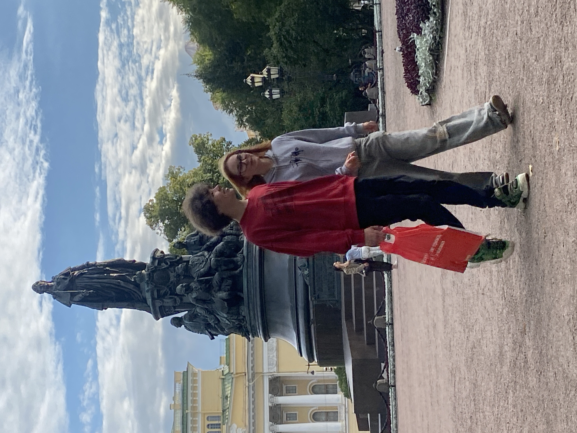 In Saint Petersburg, Russia, on a paved plaza with gravel and decorative plantings, a young man and woman walk side-by-side. The man, on the left, wears a red sweatshirt and dark pants, carrying a red plastic bag in his right hand. The woman, to his right, wears a grey hooded sweatshirt and light-colored, distressed pants, with glasses. Behind them, a grand monument, likely the Monument to Catherine the Great, stands tall against a cloudy blue sky, with a neoclassical building visible in the background. Other figures are present in the distance, further down the plaza.