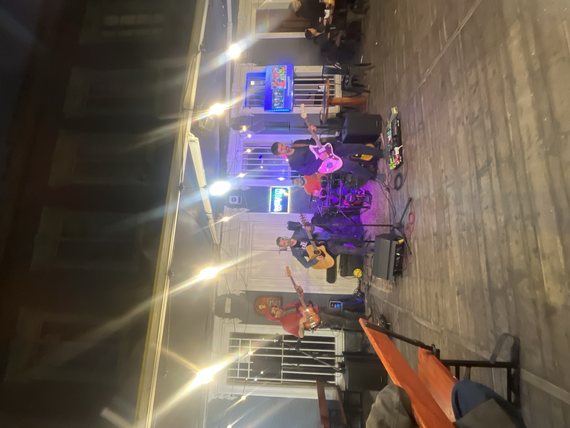 The image depicts an outdoor stage performance at night in Iquique, Chile. Four individuals are visible on stage, engaged in playing musical instruments. Two individuals are playing guitars, one is playing a bass guitar, and another is behind a drum set. The stage is set up on a wooden platform, illuminated by overhead lights and stage lighting that casts strong reflections. In the background, there's a building with windows and architectural details, suggesting an urban setting. To the right of the stage, several tables and chairs are set up, indicating an area for an audience or dining. People are seated at some of these tables, observing the performance. A television screen is mounted on the wall behind the stage, displaying images. The overall scene conveys a lively atmosphere of a live music event.