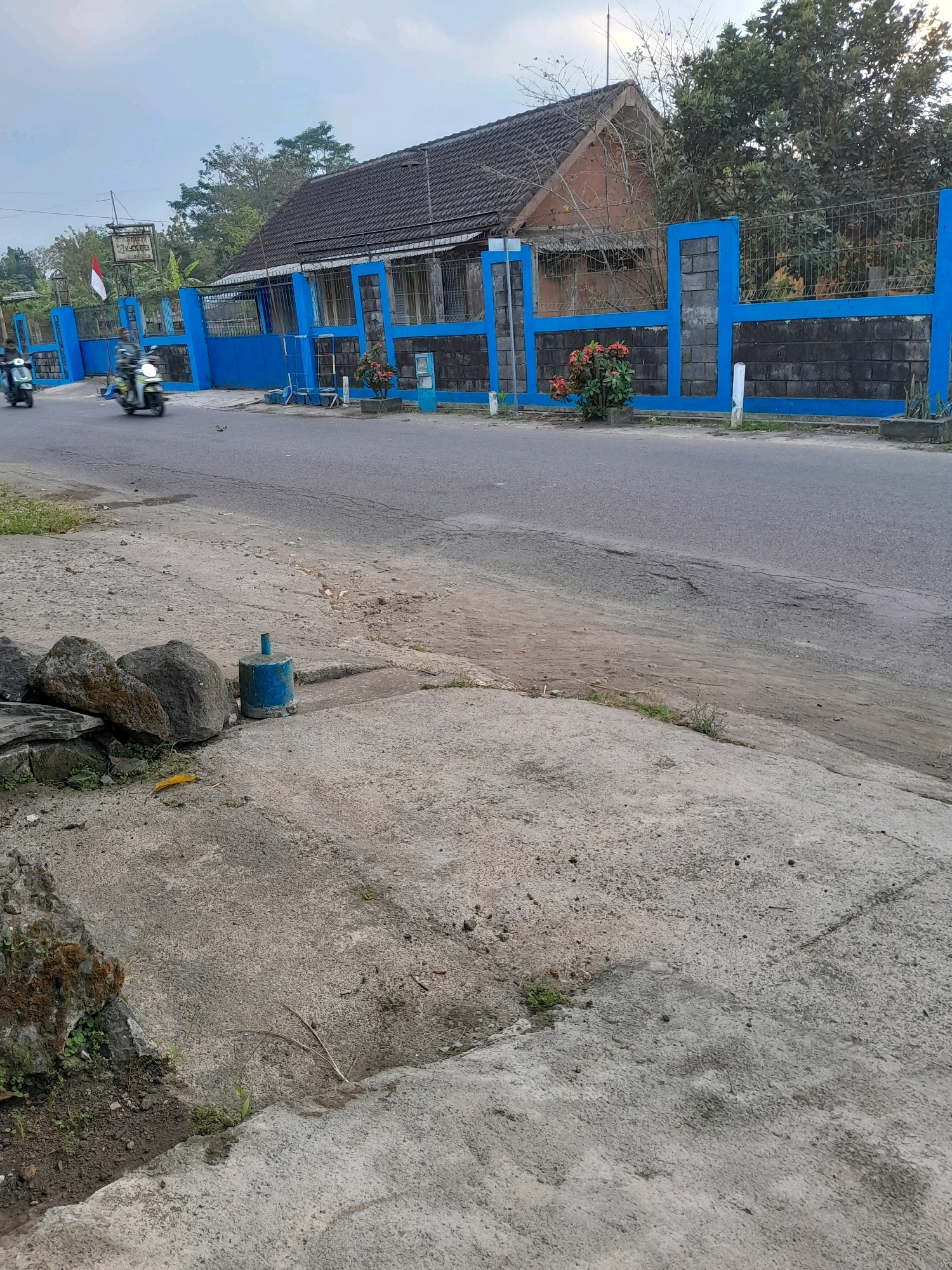 The image depicts a street scene in Glanggang, Indonesia. In the background, a building with a dark tiled roof and brick facade is visible, bordered by a blue and grey brick wall with metal fencing. A sign is mounted on a post near the entrance, and an Indonesian flag is attached to it. Two individuals on scooters are present on the paved road. In the foreground, the side of the road is made of concrete and dirt, with several large rocks and a blue cylindrical object. The sky is overcast.