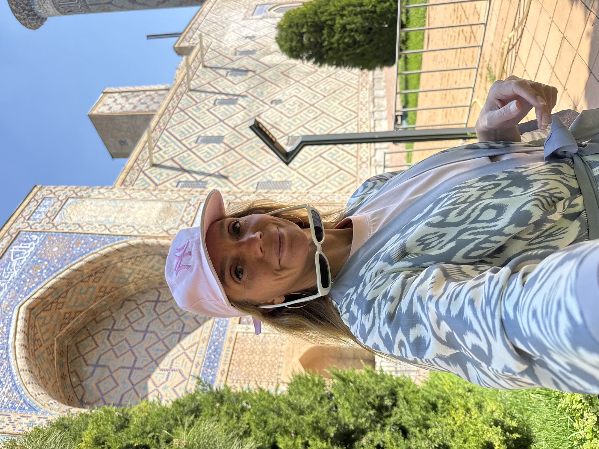 A woman wearing a pink baseball cap, white sunglasses, a light pink t-shirt, and a blue and white patterned garment is taking a selfie in Samarkand City, Uzbekistan. The background consists of detailed tilework, a section of a minaret, and greenery.