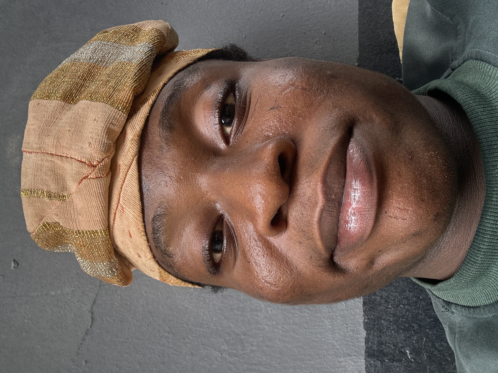 A close-up, portrait-style photograph depicts a person with dark skin, wearing a head covering that appears to be made of beige and gold fabric. The person is also wearing a green, collared shirt. The background features a gray surface and a hint of beige. The location is Unknown, Unknown.