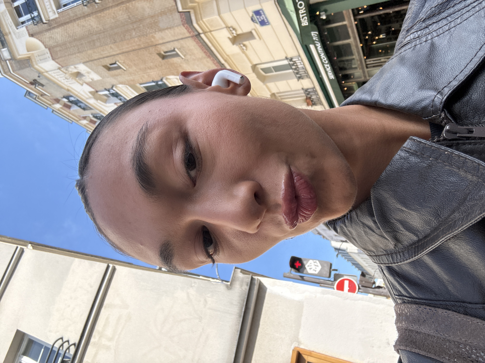 A selfie of a person wearing a black leather jacket and a white wireless earbud in their left ear, taken at street level in Paris, France. The person's face is tilted, showing a view from below and slightly to the right. In the background, there is a building with a "Bistro" sign and "Service Continu 7/7" visible, indicating a restaurant open all week. Also, the street signs in blue-and-white French style. There is a traffic light showing red. The sky is blue.