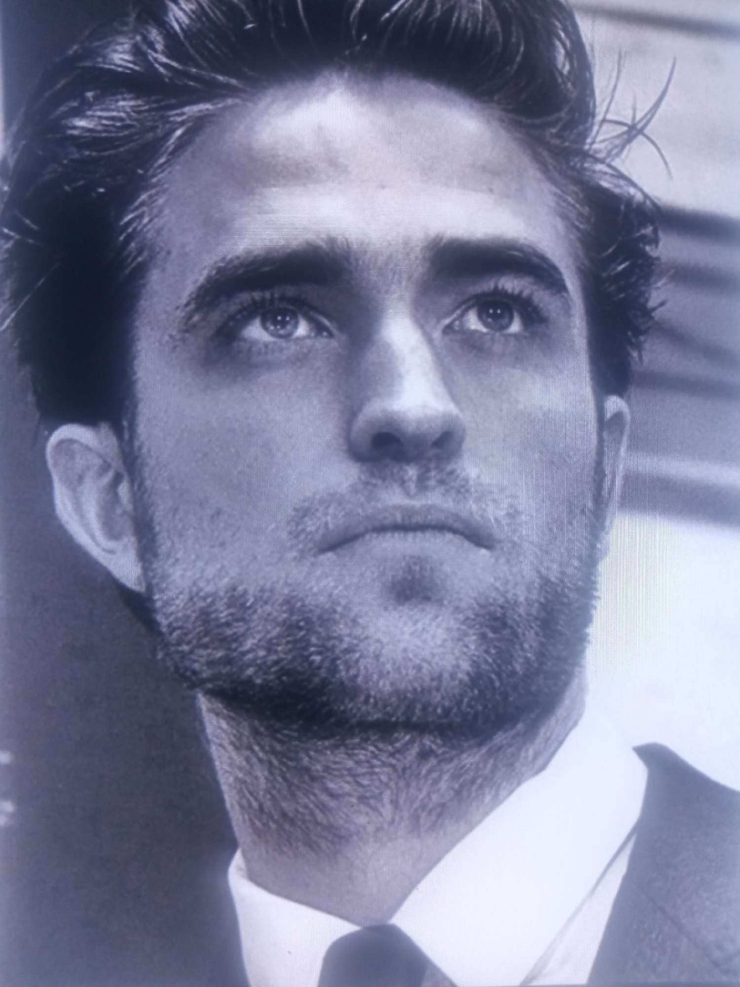 The image is a black and white, medium shot of a man's face. He is looking upwards and slightly to his left. He has dark, tousled hair, prominent eyebrows, and appears to be in his late 20s or early 30s. He has a light beard and is wearing a white collared shirt and a dark suit jacket. The background is out of focus but appears to be an indoor setting. There are no discernible landmarks or contextual clues to suggest a specific location within a city.
