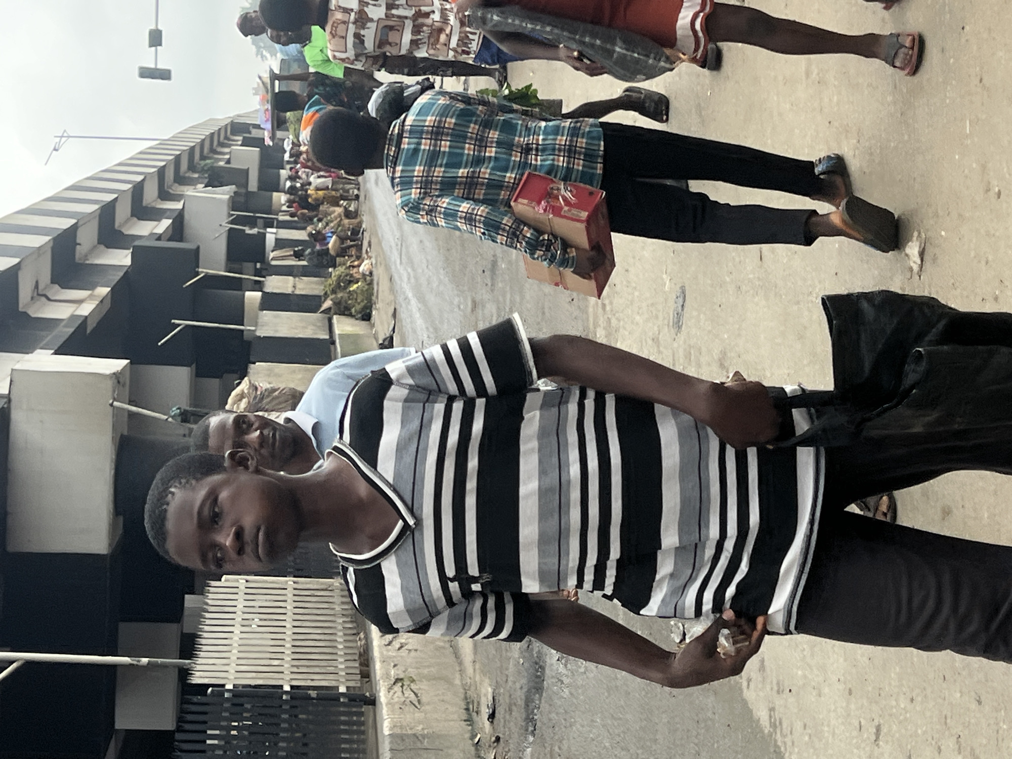 The image depicts a street scene in Nkpor, Nigeria, under an overpass. In the foreground, a young Black male is walking forward, facing slightly left, and looking ahead with a neutral expression. He is wearing a horizontally striped t-shirt with black, white, and gray bands, and black trousers. He is carrying a black cloth bag in his right hand and holding a small, light-colored object in his left hand. Behind him, another Black male is partially visible, wearing a light blue collared shirt. Further back, a group of people are seen congregated along the side of the street, some appearing to be engaged in commerce, with stalls and goods displayed. The ground is paved and appears somewhat littered. The overpass structure dominates the background, with concrete pillars and a road surface visible above. The overall atmosphere suggests a busy, outdoor market or thoroughfare in an urban environment.
