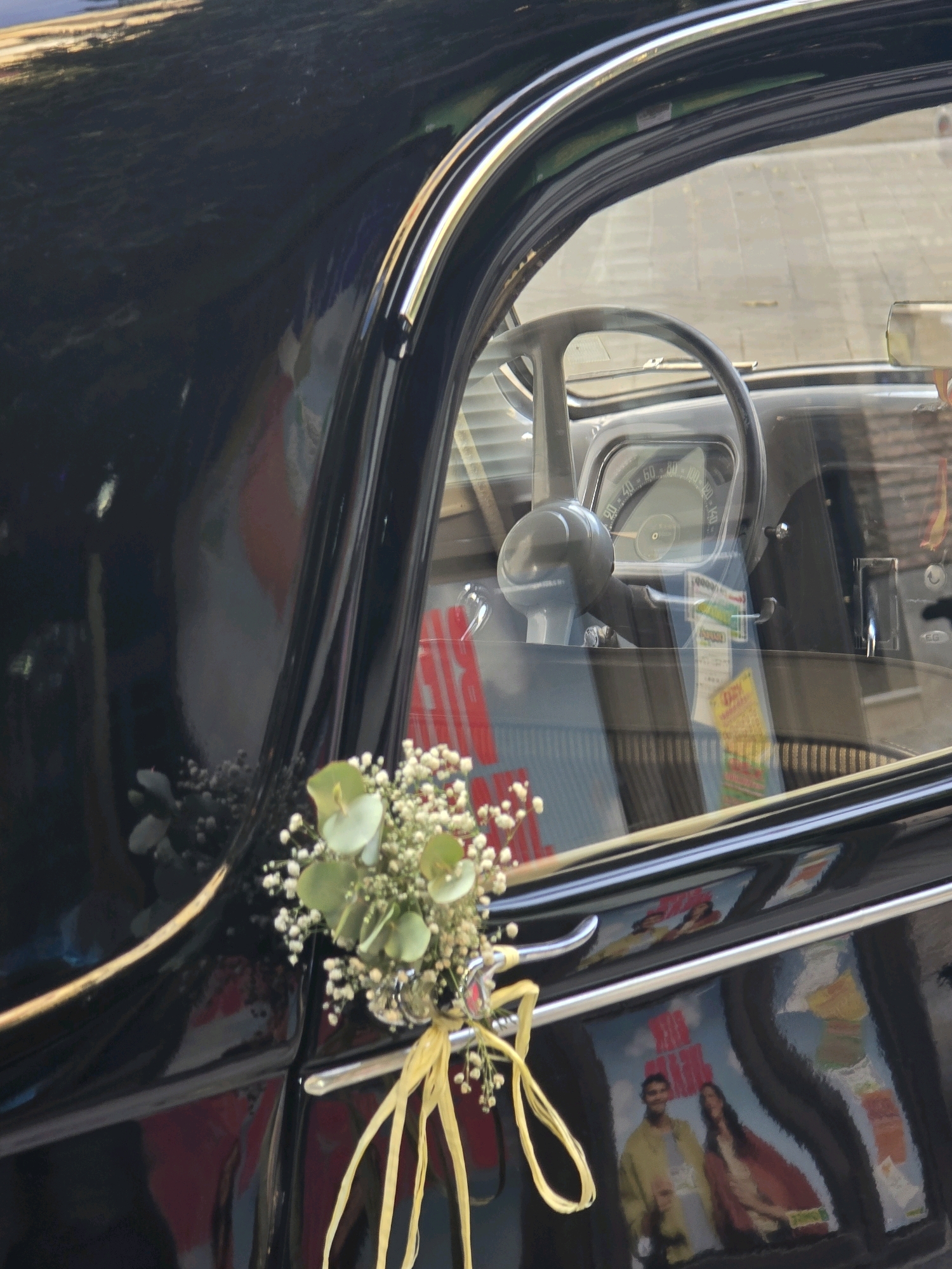 The image is a close-up shot of the side of a black vintage car, possibly from the 1950s or 1960s, parked outdoors. The car has a shiny, reflective surface, showing reflections of the surroundings. A small bouquet of white baby's breath and green eucalyptus leaves, tied with a yellow ribbon, is attached to the car's door handle. Through the car's window, the steering wheel and dashboard of the interior are visible. Reflections in the car's paint and window suggest the car is parked on a street with buildings and possibly trees. There are posters or advertisements reflected on the car door, one showing a couple. The light suggests it is daytime. The overall impression is that the car is decorated for an event like a wedding or celebration. While the image is taken in Granada, Spain, there are no specific landmarks or visual cues in the image that definitively place it in Granada.