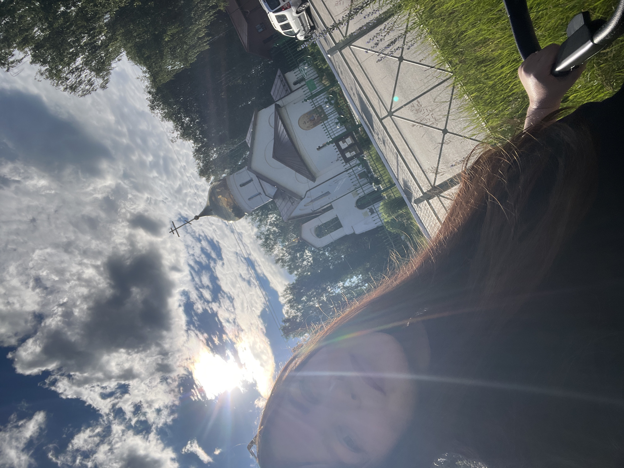 The image, taken in Noviy, Russia, shows a young woman taking a selfie outdoors. She is in the foreground, with her face and long brown hair filling the left side of the frame. The background features a white church with a golden dome and a cross, surrounded by trees. The sun is bright, casting lens flares and illuminating the sky, which is filled with clouds. A metal fence and a street are also visible. A white car is parked near the church.