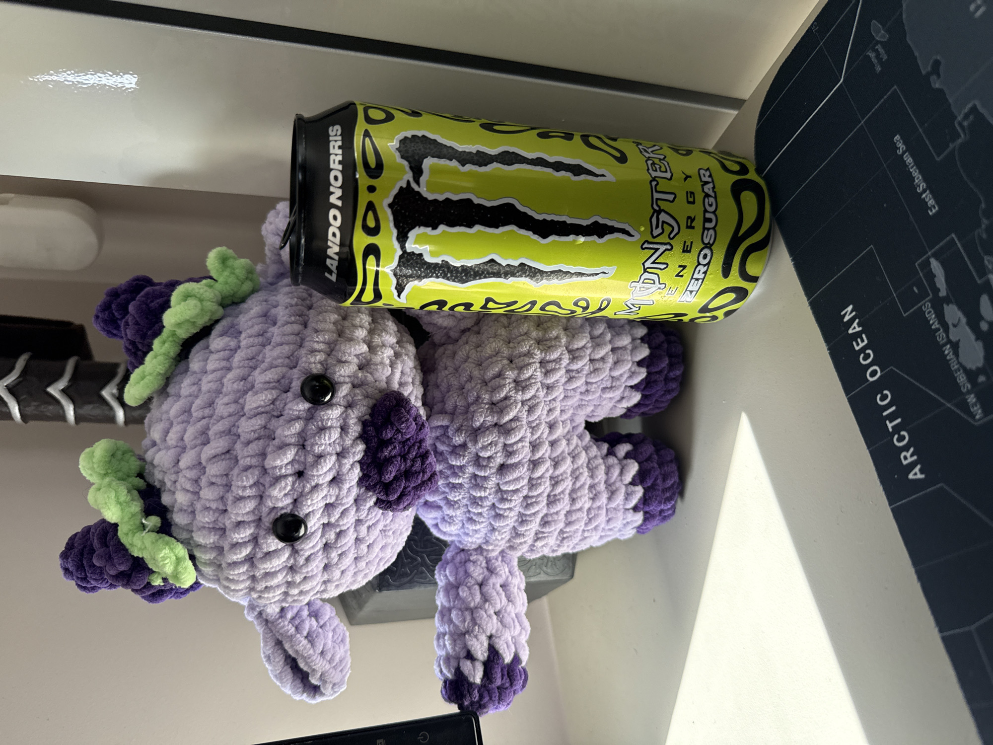 The image shows a crocheted purple unicorn toy with purple and green details next to a can of "Monster Energy Zero Sugar" drink, the Lando Norris edition. The unicorn toy has black button eyes and a mane and tail made of dark purple and bright green yarn. The can is yellow with black claw marks and the "Monster" logo prominently displayed. The can is labeled "Lando Norris" near the top. The objects are positioned on a white surface. A portion of a dark blue mat or pad with a map is visible on the right, with the words "ARCTIC OCEAN" and "East Siberian Sea". The photo was taken from above. There is no information to identify the location as Sofia, Bulgaria.