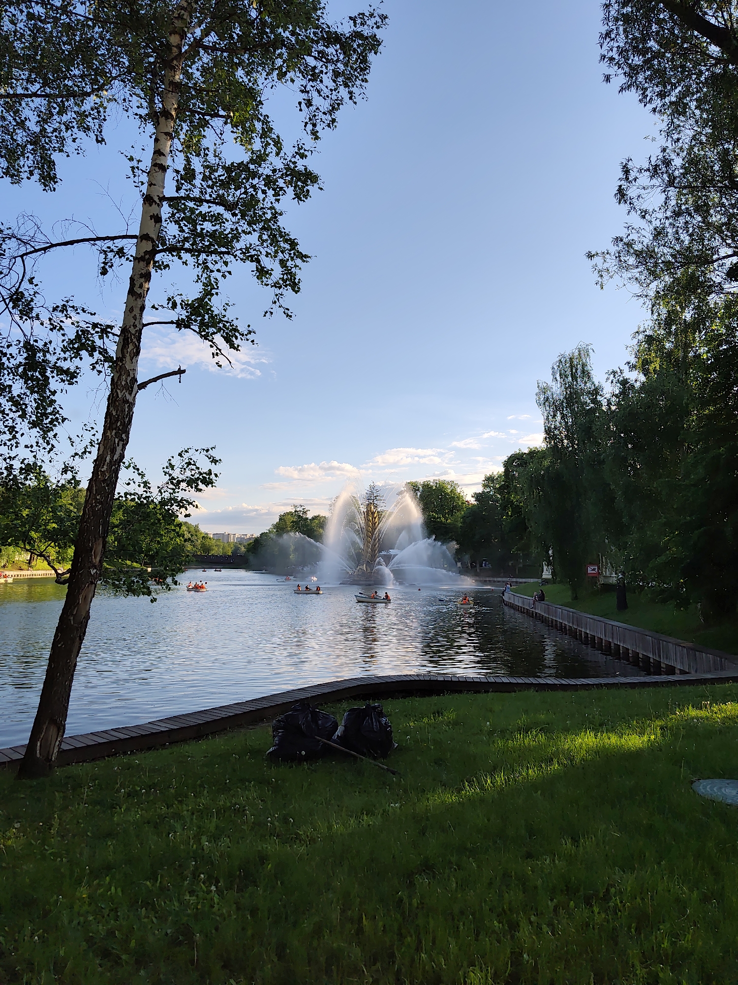 A water landscape in Moscow, Russia, features a fountain at the center, surrounded by a body of water dotted with several small boats, each carrying multiple people. Along the water's edge, there is a wooden walkway adjacent to a grassy area. In the foreground, bags are located on the grass near the water. The sky is blue with some clouds. Trees frame the scene on both sides. A birch tree stands on the left.