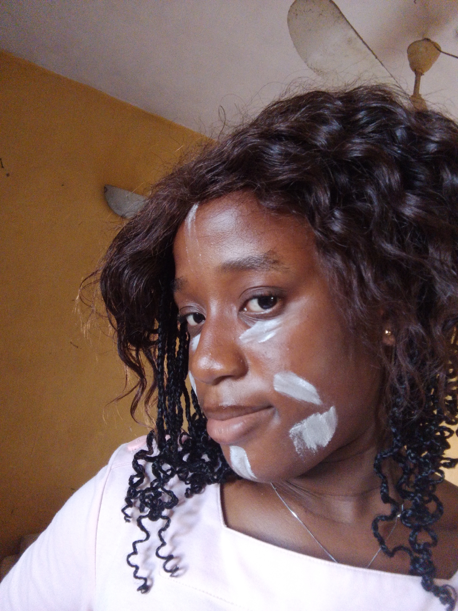 The image shows a young Black woman with dark, curly hair and braids. She is wearing a light pink shirt and a thin silver necklace. Her face is partially covered with white cream or paint, applied in a decorative pattern on her cheeks and under her eyes. She has a gold earring in her visible ear. The background is a textured, yellowish wall and a portion of a ceiling fan. The overall lighting suggests an indoor setting, possibly a room in Umuahia, Nigeria, given the context. The woman is looking directly at the camera, and her expression is neutral.