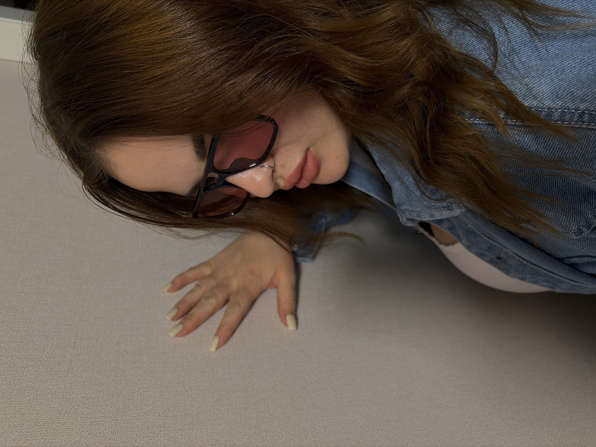 A woman with auburn hair, wearing a denim jacket and pink tinted glasses, rests her hand on a textured surface. She has a nose ring. Part of a white garment, possibly a bra or tank top, is visible beneath her jacket. The location cannot be determined as Krasnodar, Russia, from the provided image.