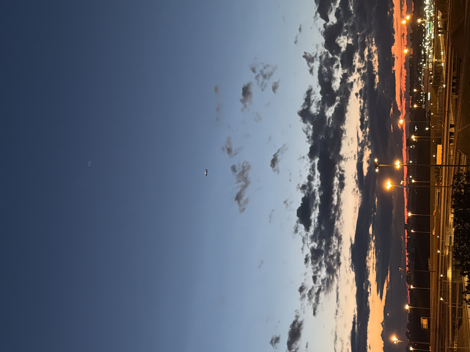 The image captures an aerial view of a cityscape at dusk. The sky transitions from a deep blue at the top to hues of orange and red near the horizon, indicating the setting sun. Dark, textured clouds are scattered across the sky, some illuminated by the fading light. An airplane is visible in the upper portion of the sky, a small silhouetted object against the vast expanse. In the foreground, the cityscape is dotted with streetlights casting a warm glow. Multiple roads and overpasses are visible, suggesting a busy urban area. The overall scene depicts a twilight ambiance over a city.